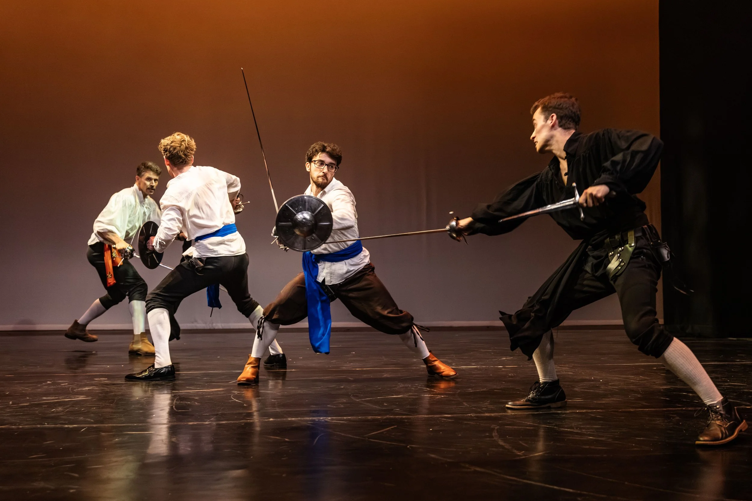 Four men in historical costumes engaging in a staged fencing duel on a dark stage.