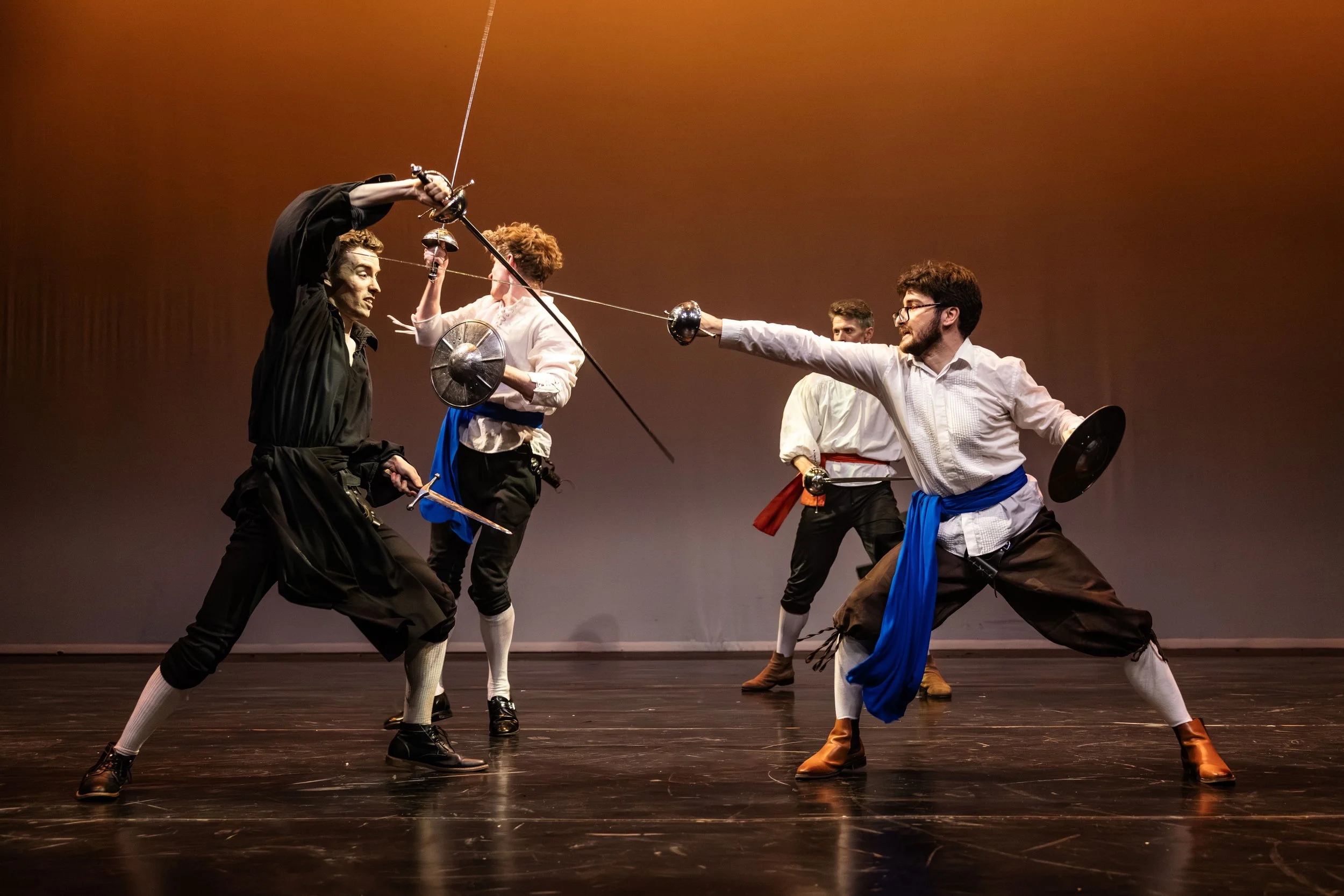 Four performers dressed in historical costumes engaged in a staged fencing duel on stage, with a plain background.