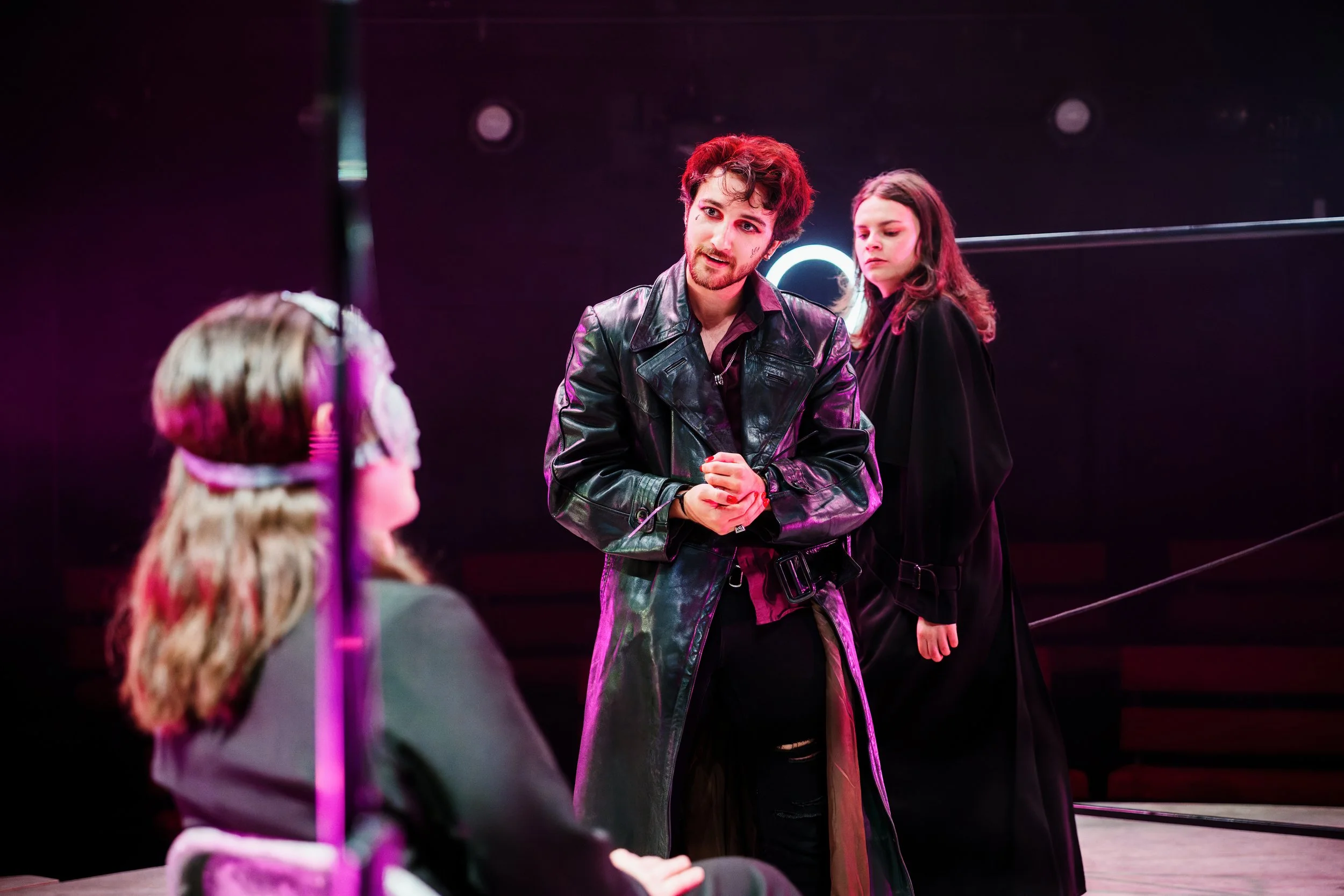 Theater scene with three actors: a young woman in a wheelchair with brown hair, a young man with curly hair in a leather coat, and a young woman with straight hair in a black dress, on stage with dark background and neon lights.