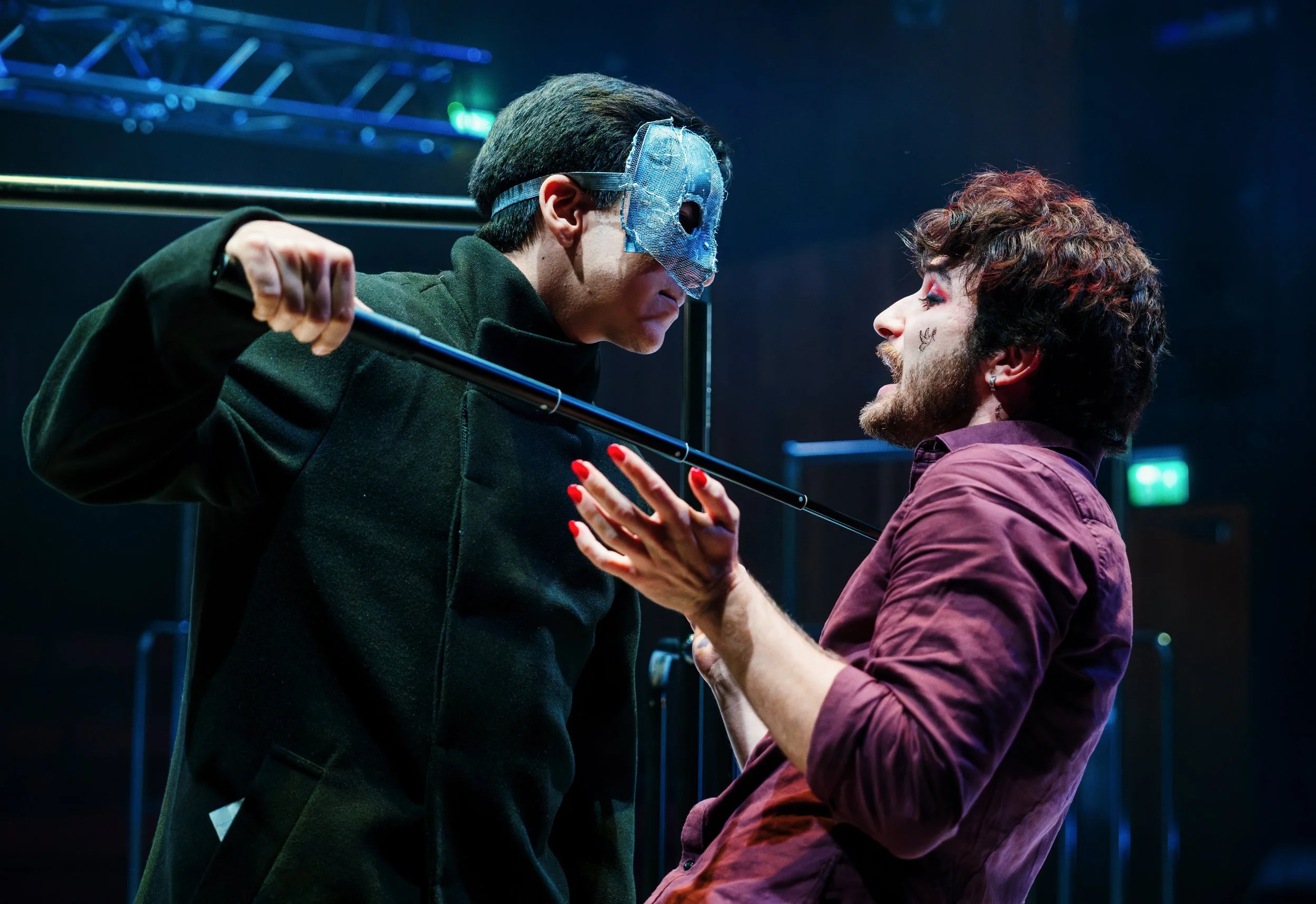 Two men engaged in a theatrical confrontation, one wearing a blue mask and black jacket, holding a cane, and the other with curly hair and a beard, wearing a purple shirt, on a stage with blue lighting.