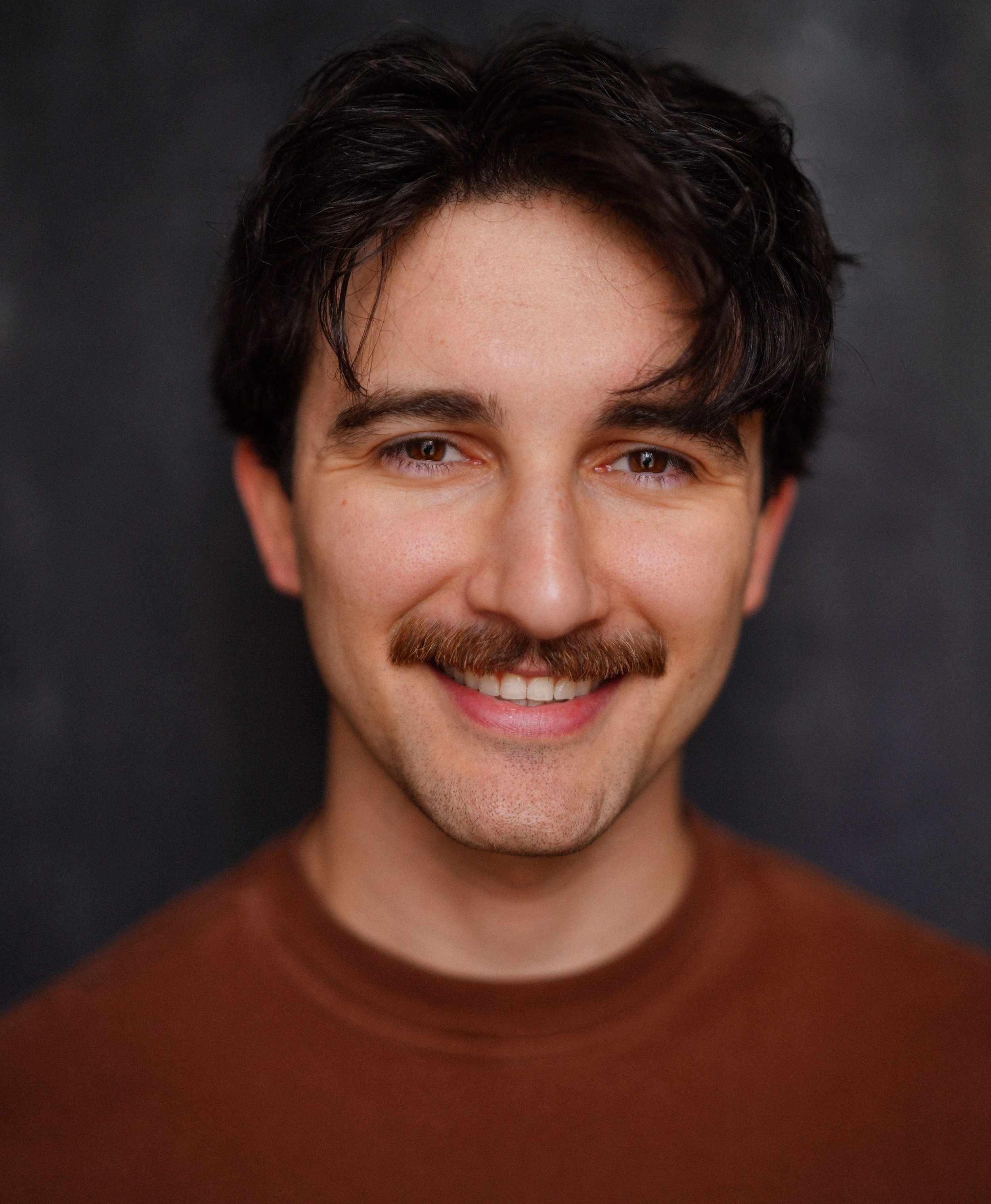 A young man with dark wavy hair, a mustache, and a friendly smile, wearing a brown shirt against a dark background.