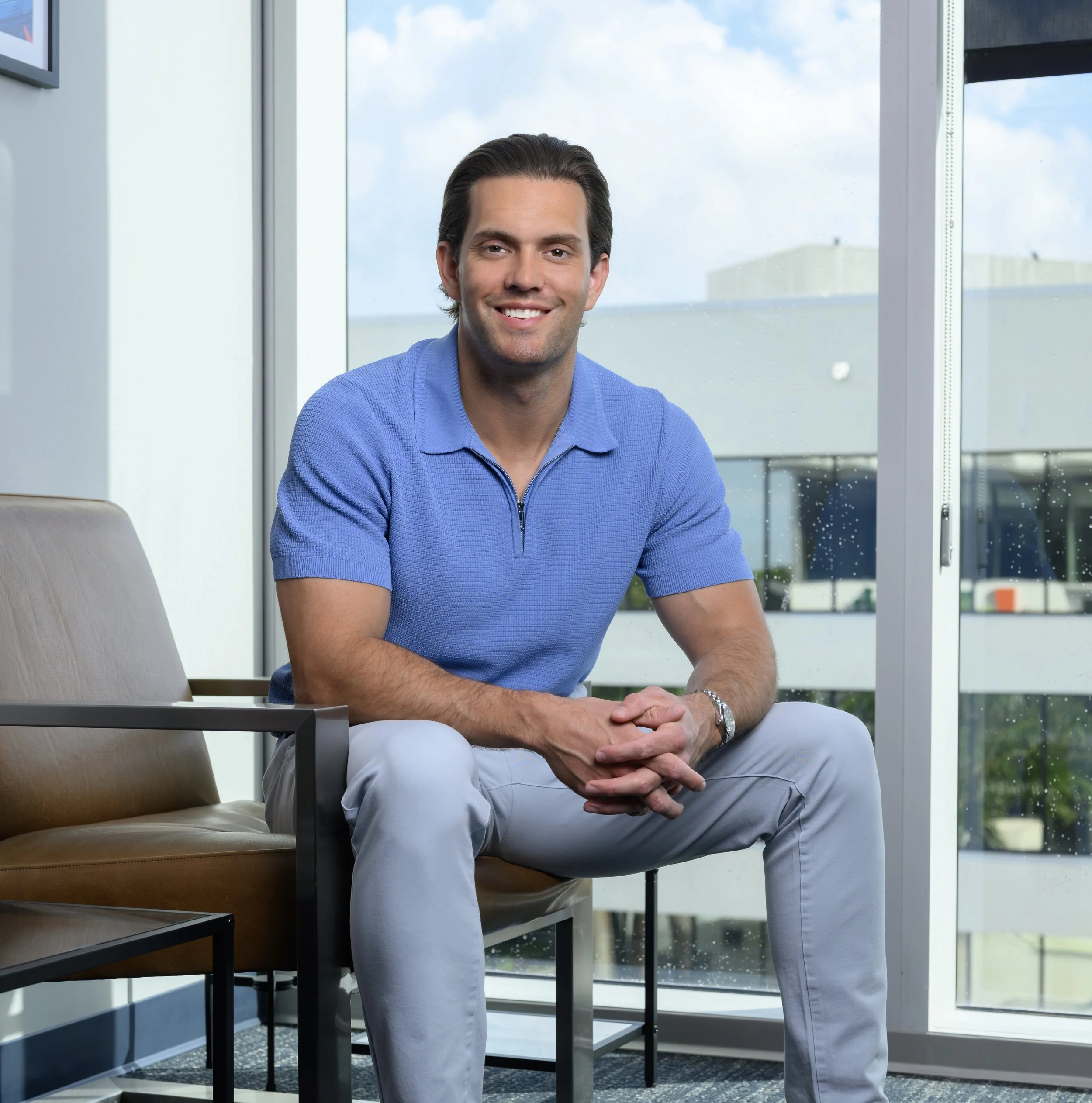 A man with dark hair and a smile, wearing a blue zip-up sweater and white pants, sitting with his hands clasped in front of him in a modern office setting with large windows and a cityscape background.