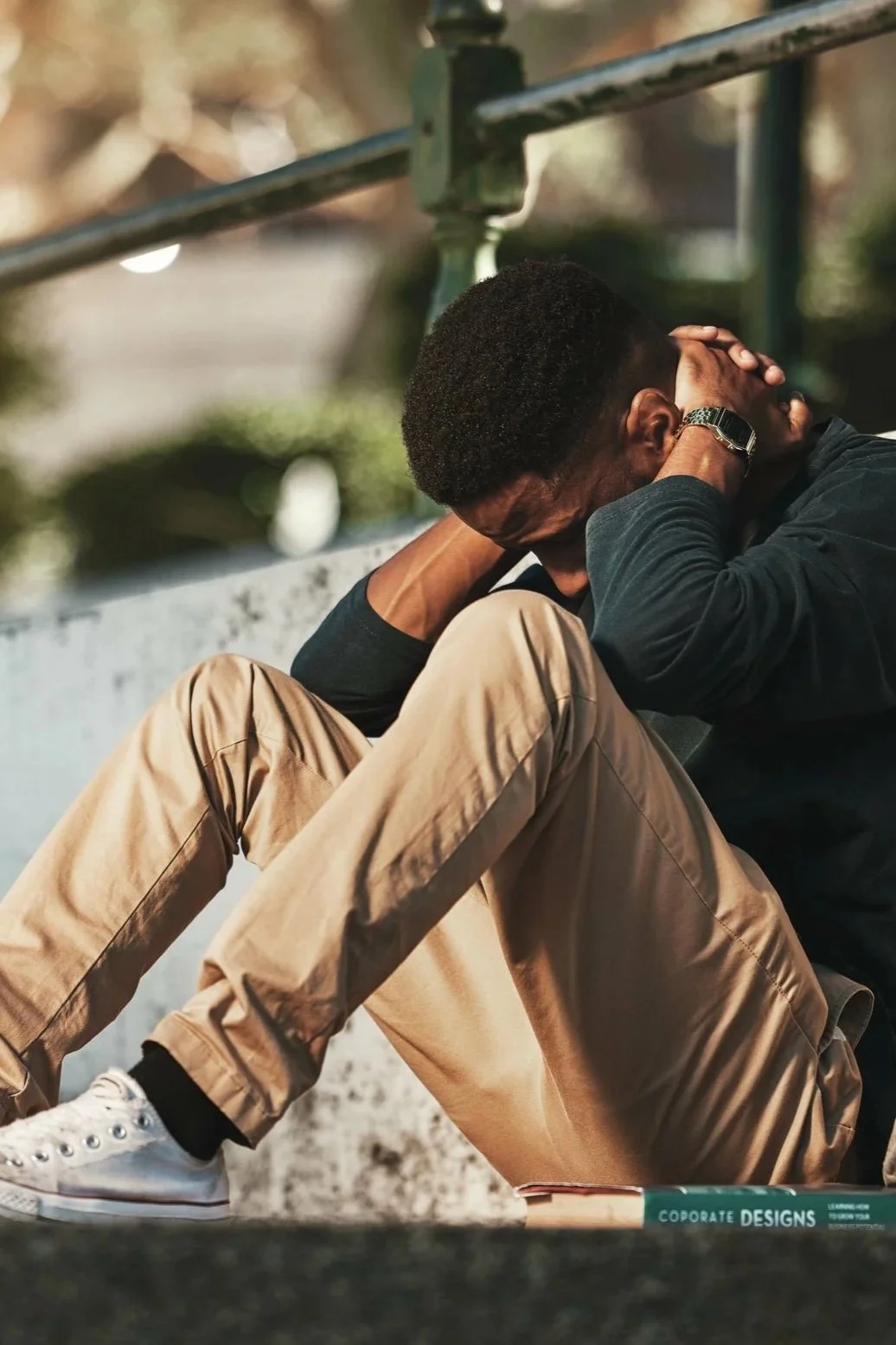 A young man sitting on a step outdoors, holding his head in his hands, appearing distressed or upset.