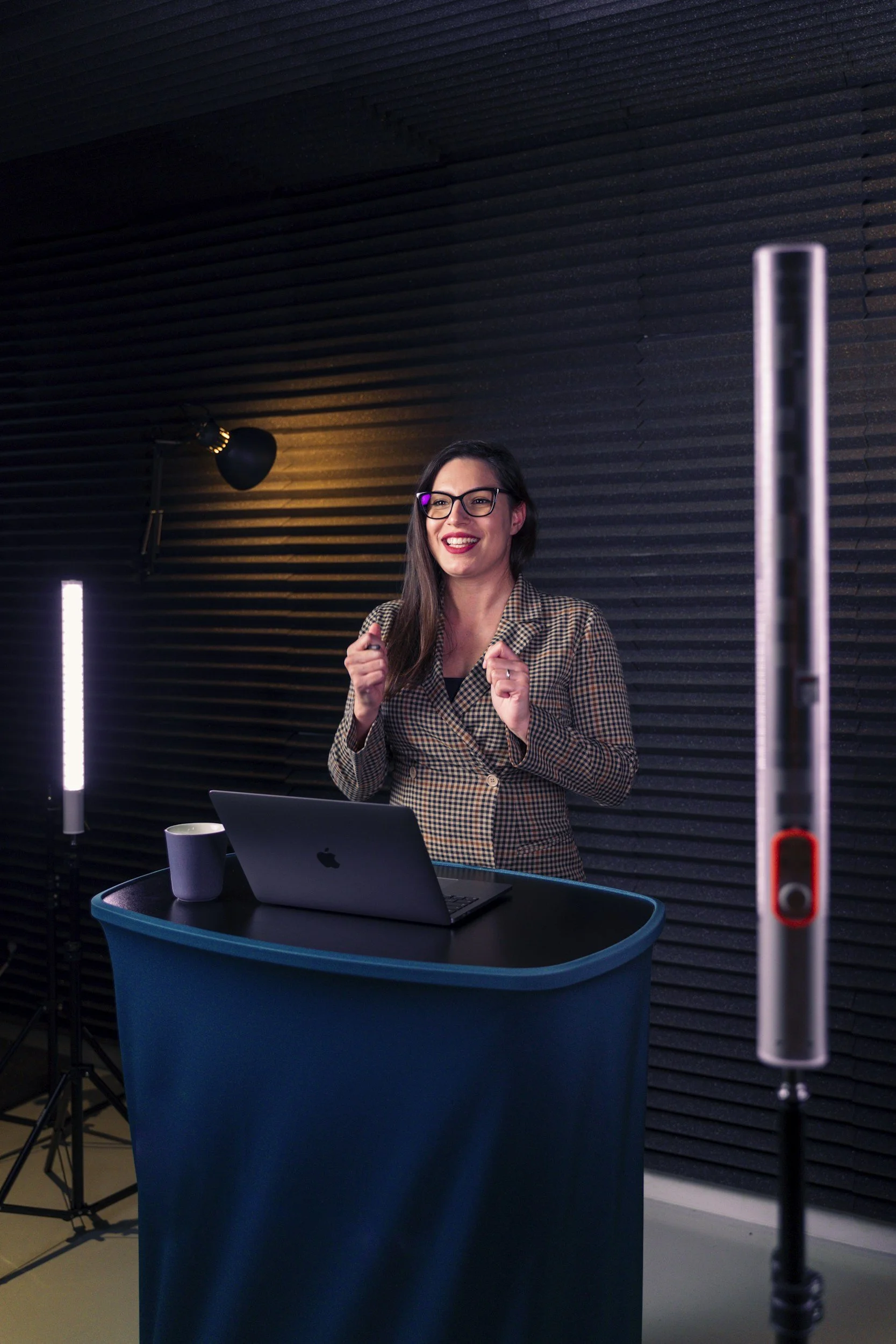 A woman with glasses and long dark hair, smiling and gesturing with her hands, standing behind a blue podium in a dimly lit room with black soundproof wall panels, a laptop, a cup, and a lighting setup.