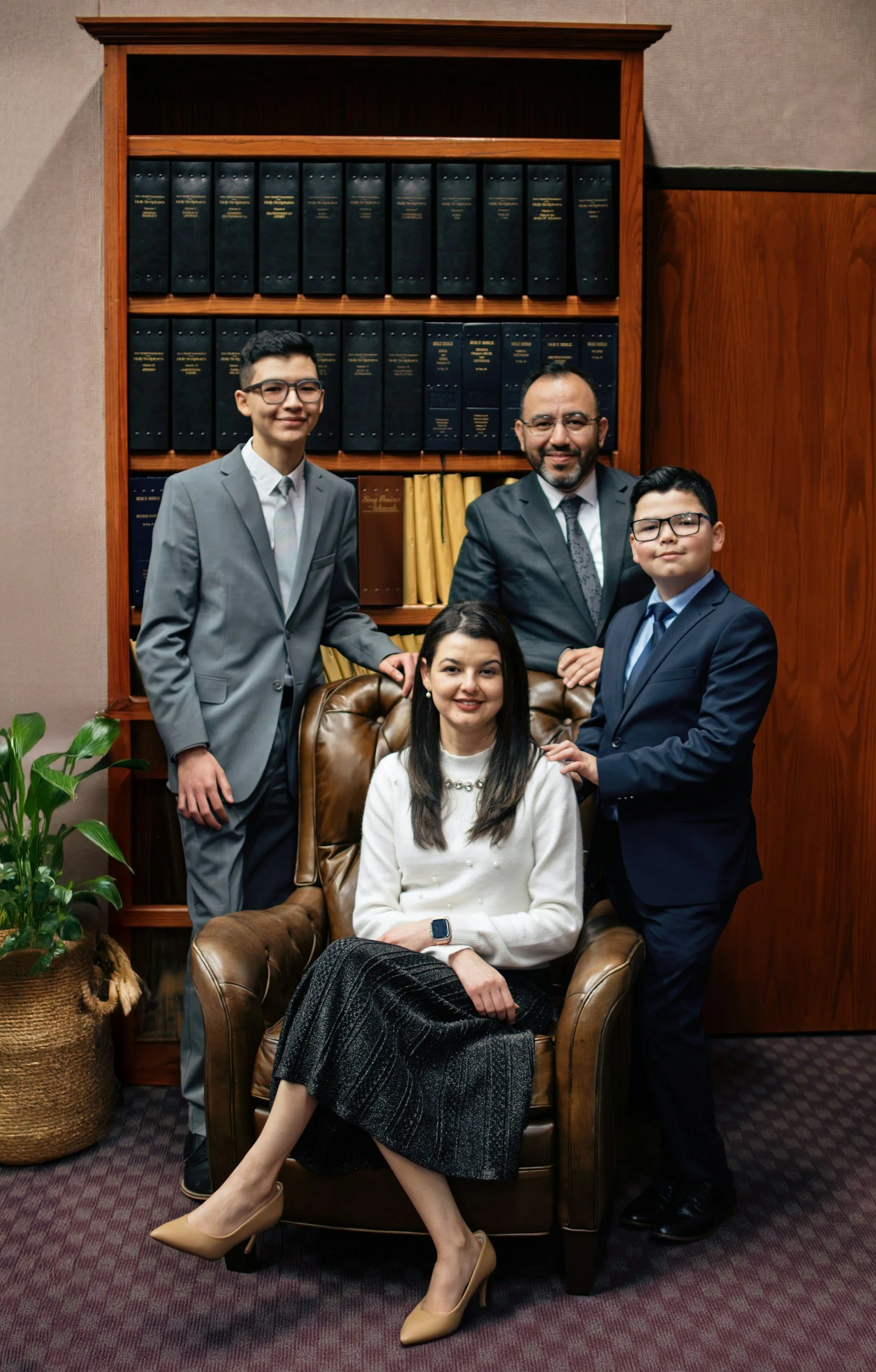 Group of five professionally dressed individuals, three men and two women, posing together in an office or conference room. One woman is seated in a leather armchair, while the others stand around her, and they are all smiling at the camera. A bookshelf with dark and yellow books and a plant are visible in the background.