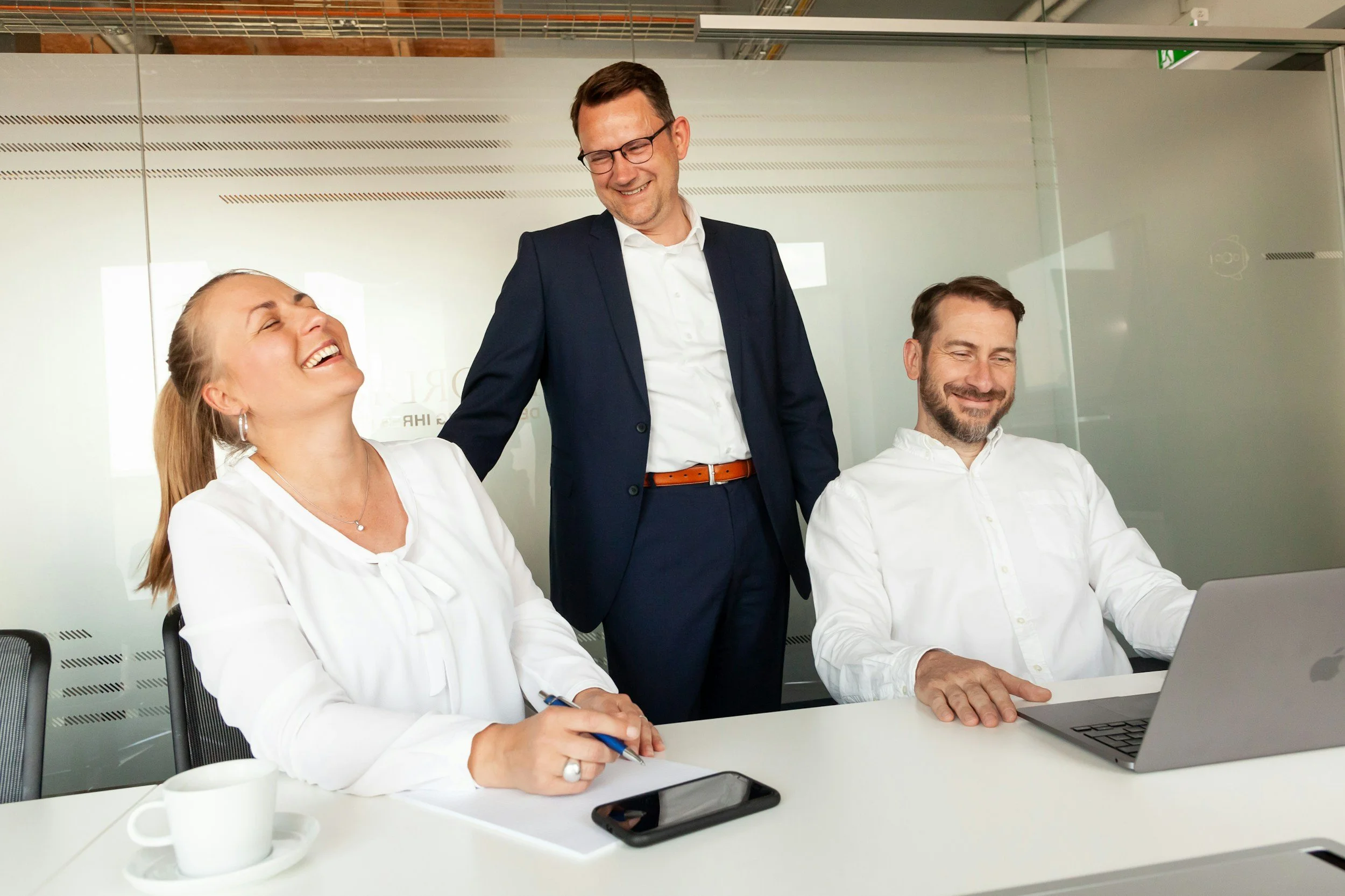 Three business professionals in a conference room, two sitting at a table with laptops and a coffee mug, one standing and smiling, engaging in a conversation or meeting.