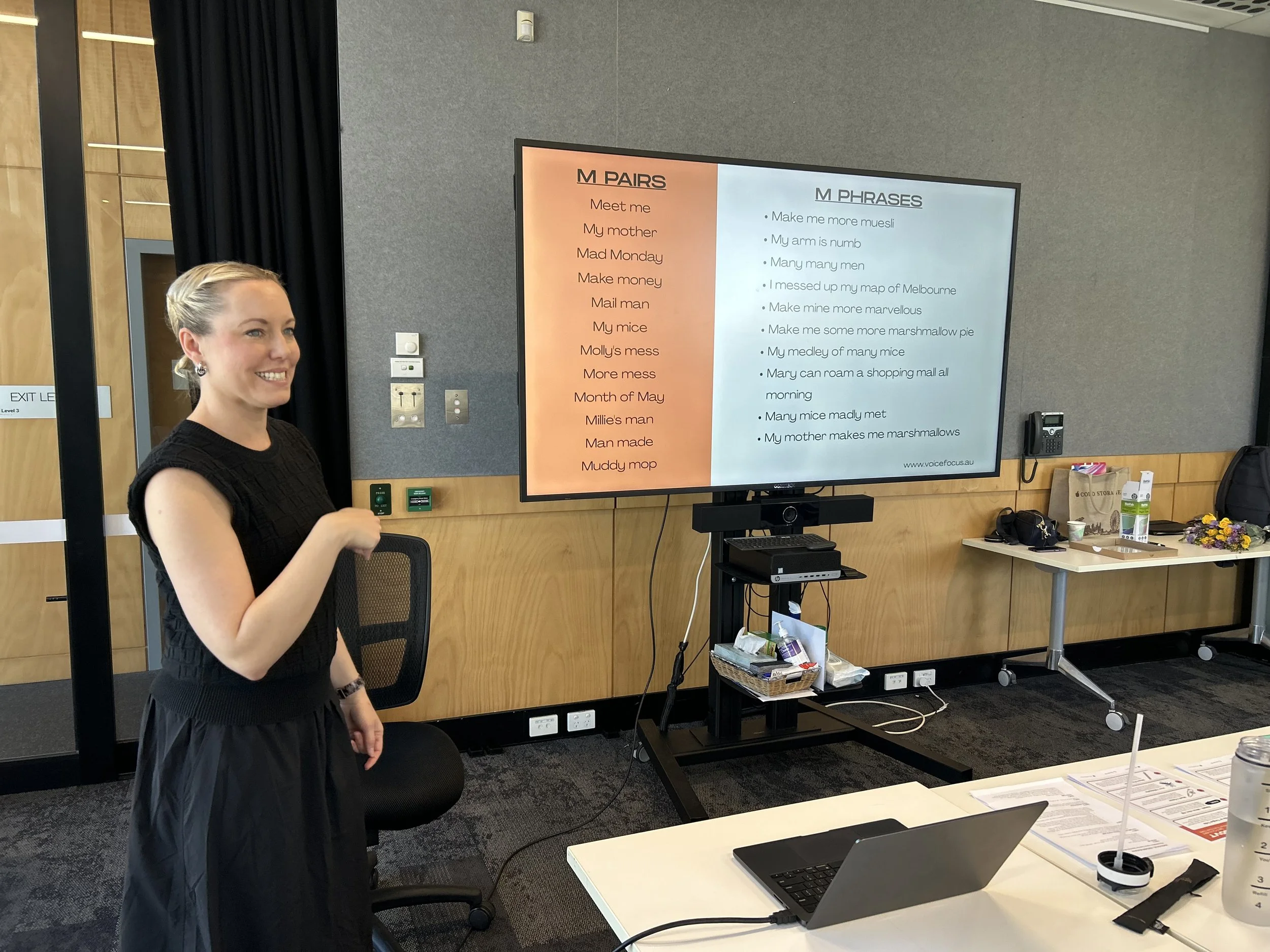 A woman smiling in front of a large screen displaying educational content titled 'M PAIRS' and 'M PHRASES' in a classroom or conference room.