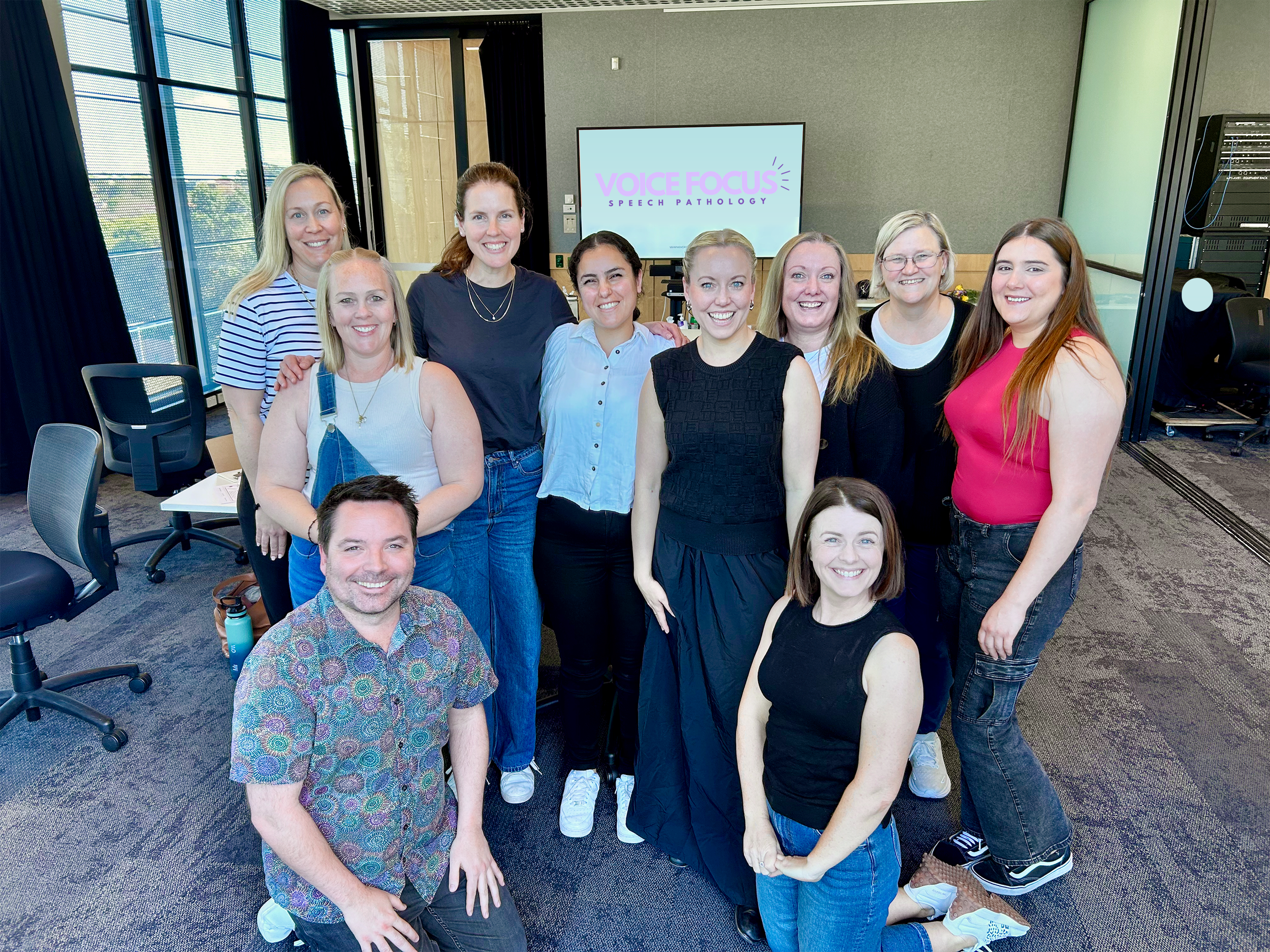 Group of nine women posing together in a room with large windows, a television displaying 'Voice Focus Speech Pathology,' and office chairs and equipment.