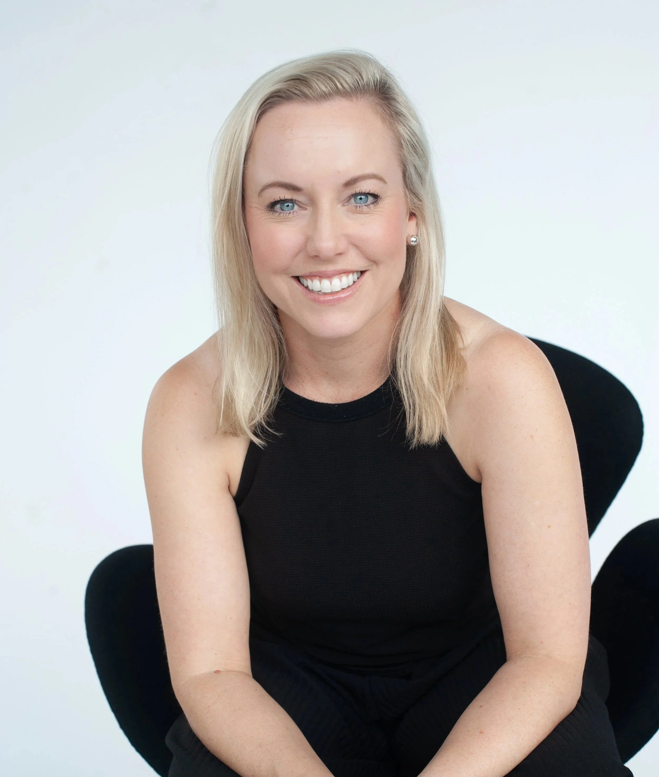 A smiling woman with long blonde hair, blue eyes, wearing pearl earrings and a black sleeveless top, sitting on a black chair against a plain white background.