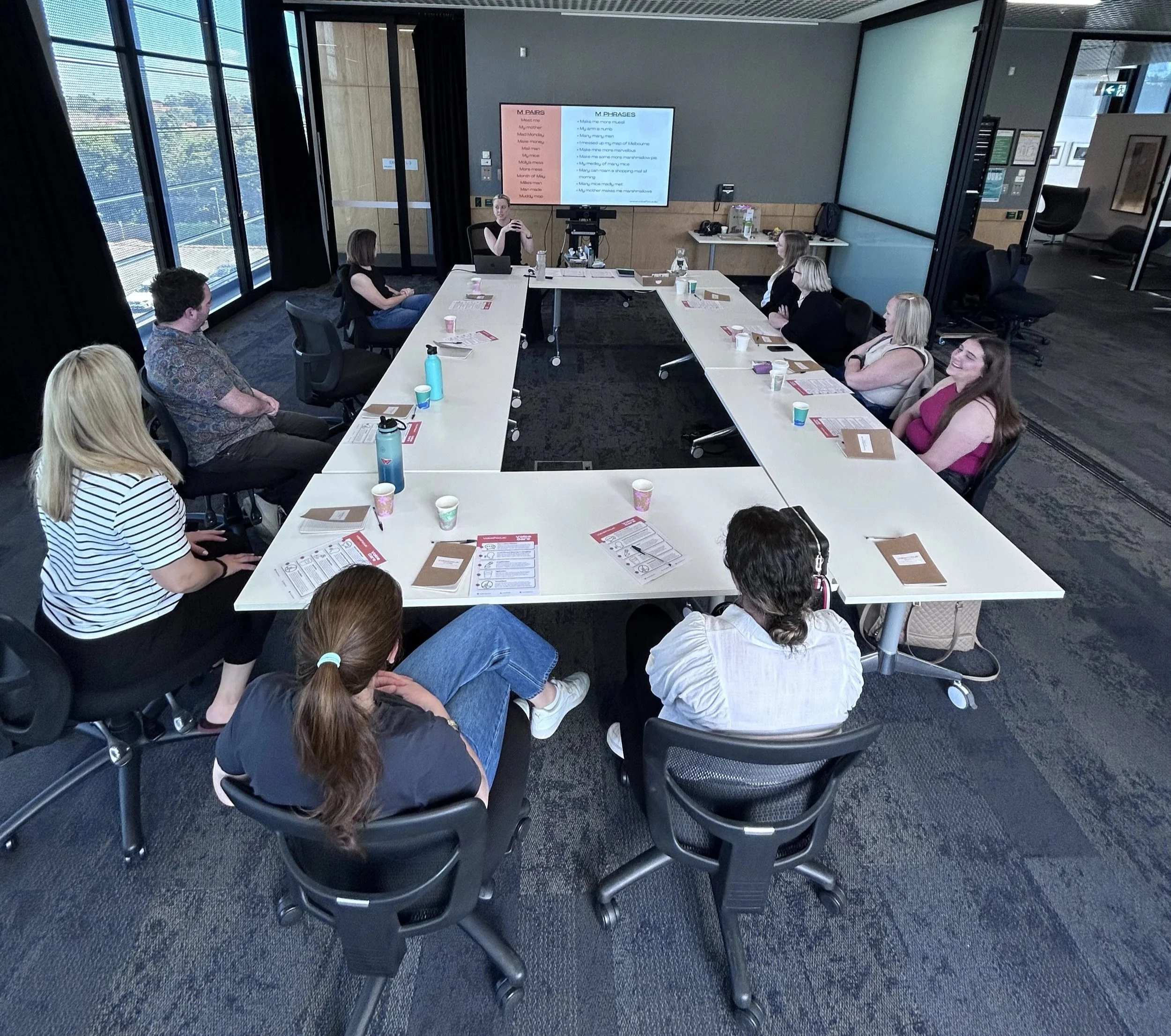 A group of people seated around a U-shaped conference table in a modern office, with a woman standing at the front near a large screen displaying a presentation.
