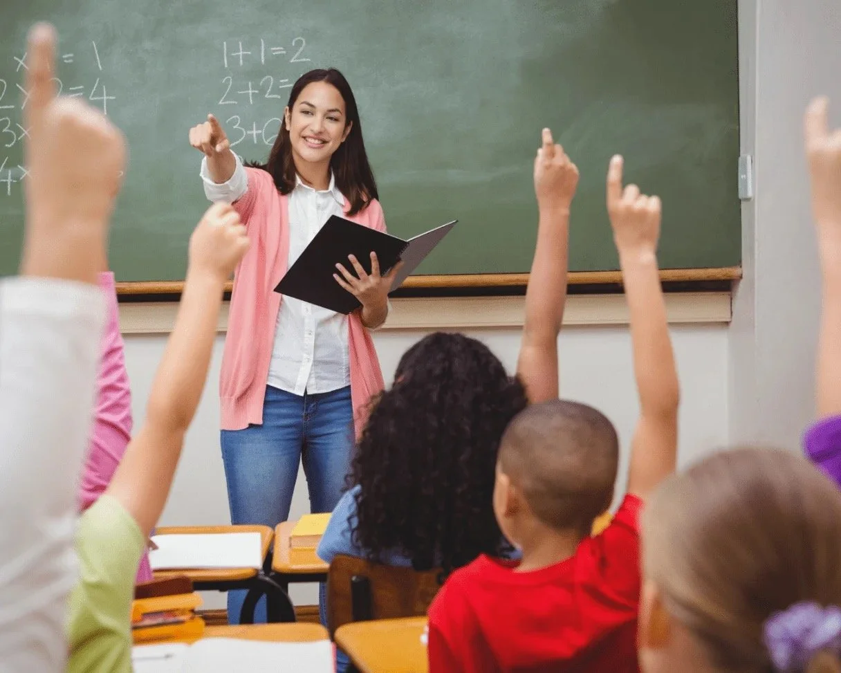 A female teacher standing at the chalkboard in a classroom, holding a book and smiling as students raise their hands to answer questions.