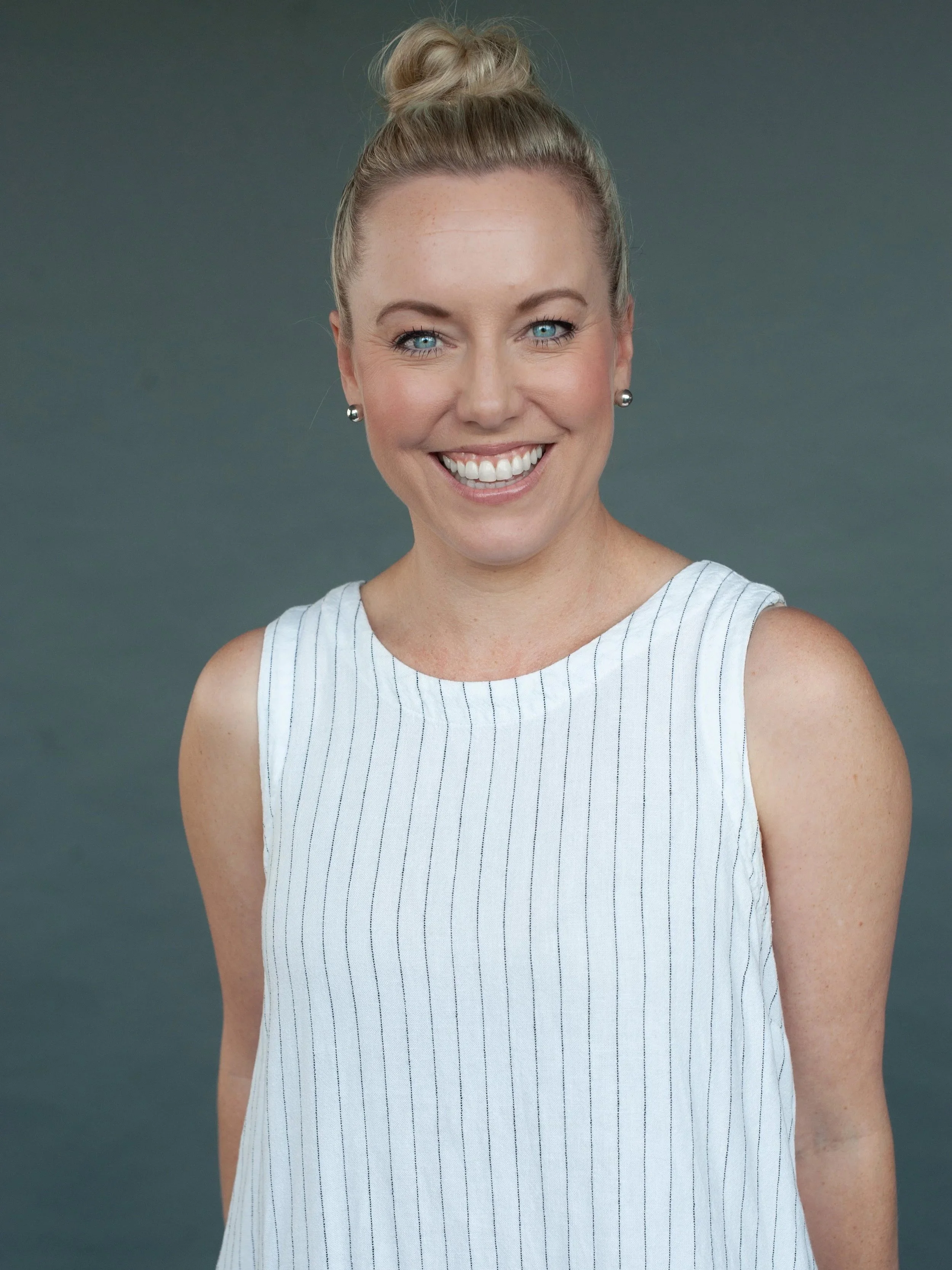 A woman with blond hair styled in a bun, smiling with blue eyes, wearing silver earrings and a sleeveless white and blue striped top, standing against a plain gray background.