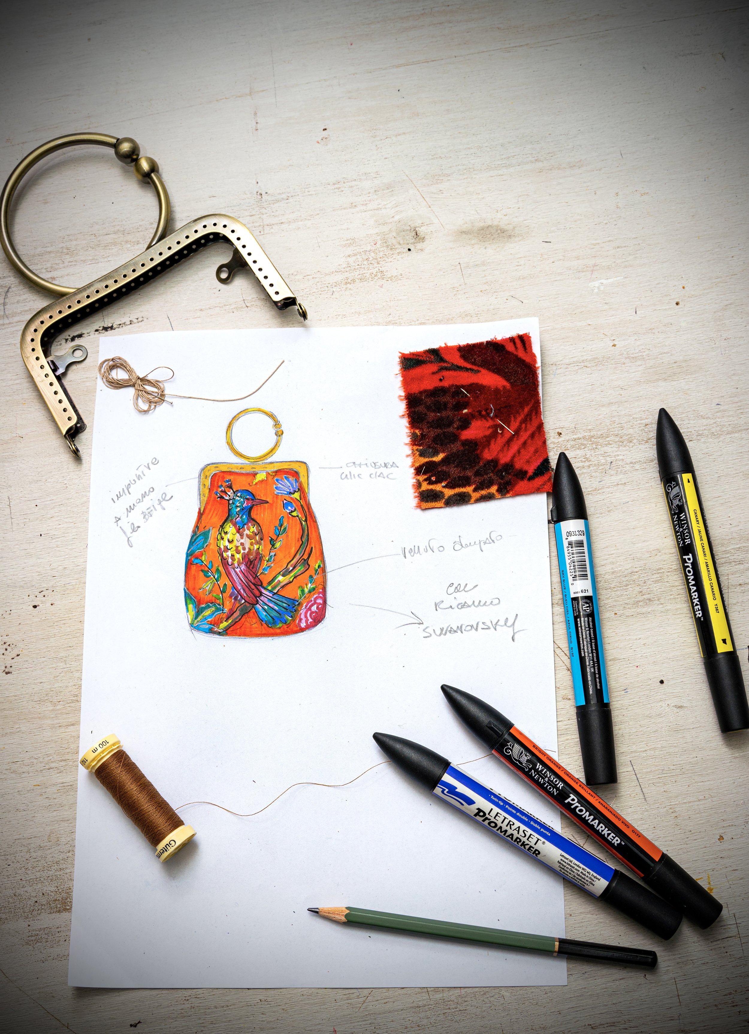 Flat lay of a design sketch of a colorful bird on a pendant, surrounded by fabric swatches, markers, thread spool, and jewelry tools on a light wooden surface.