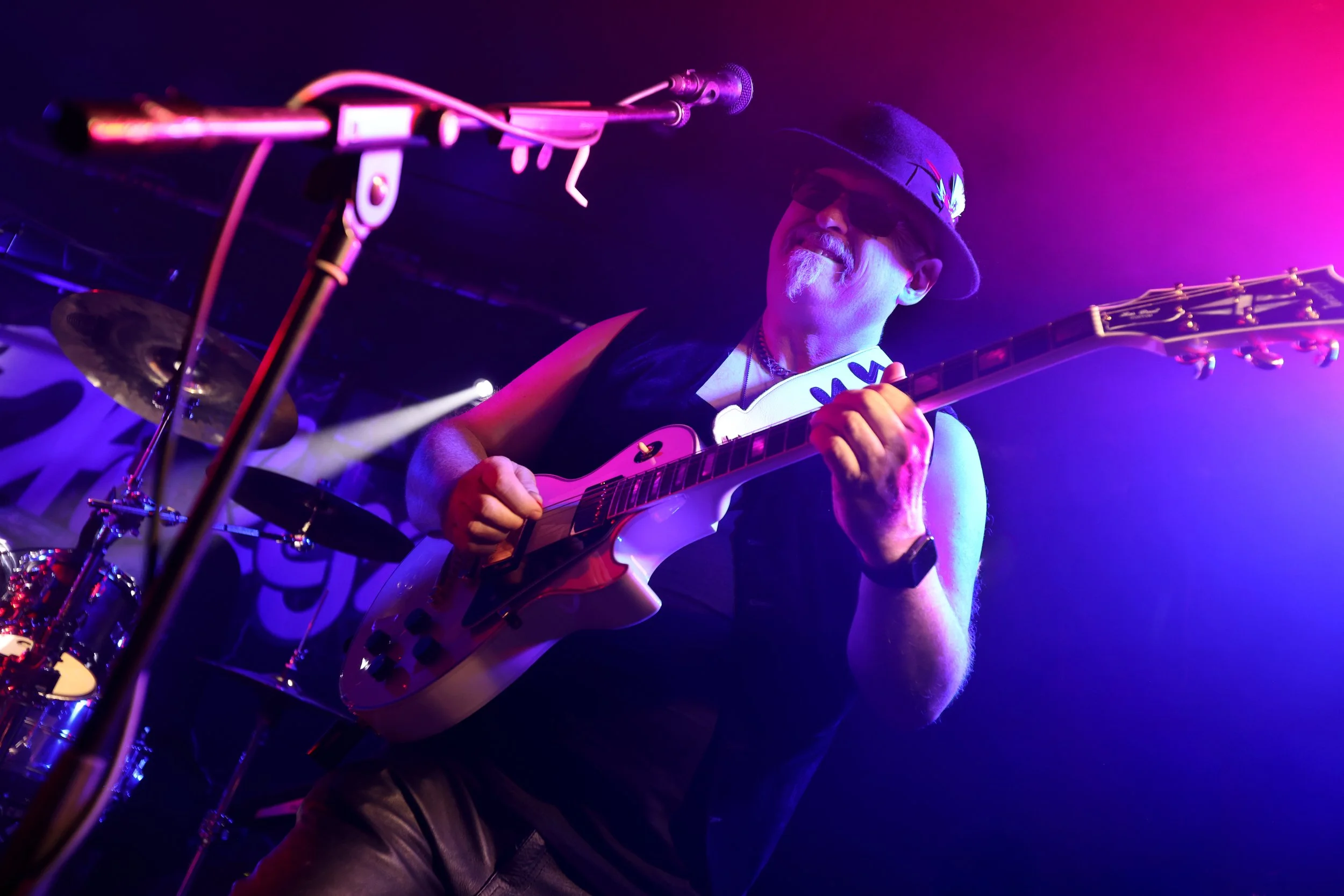 A male musician with a beard, wearing a hat, playing an electric guitar on stage with purple and pink lighting.
