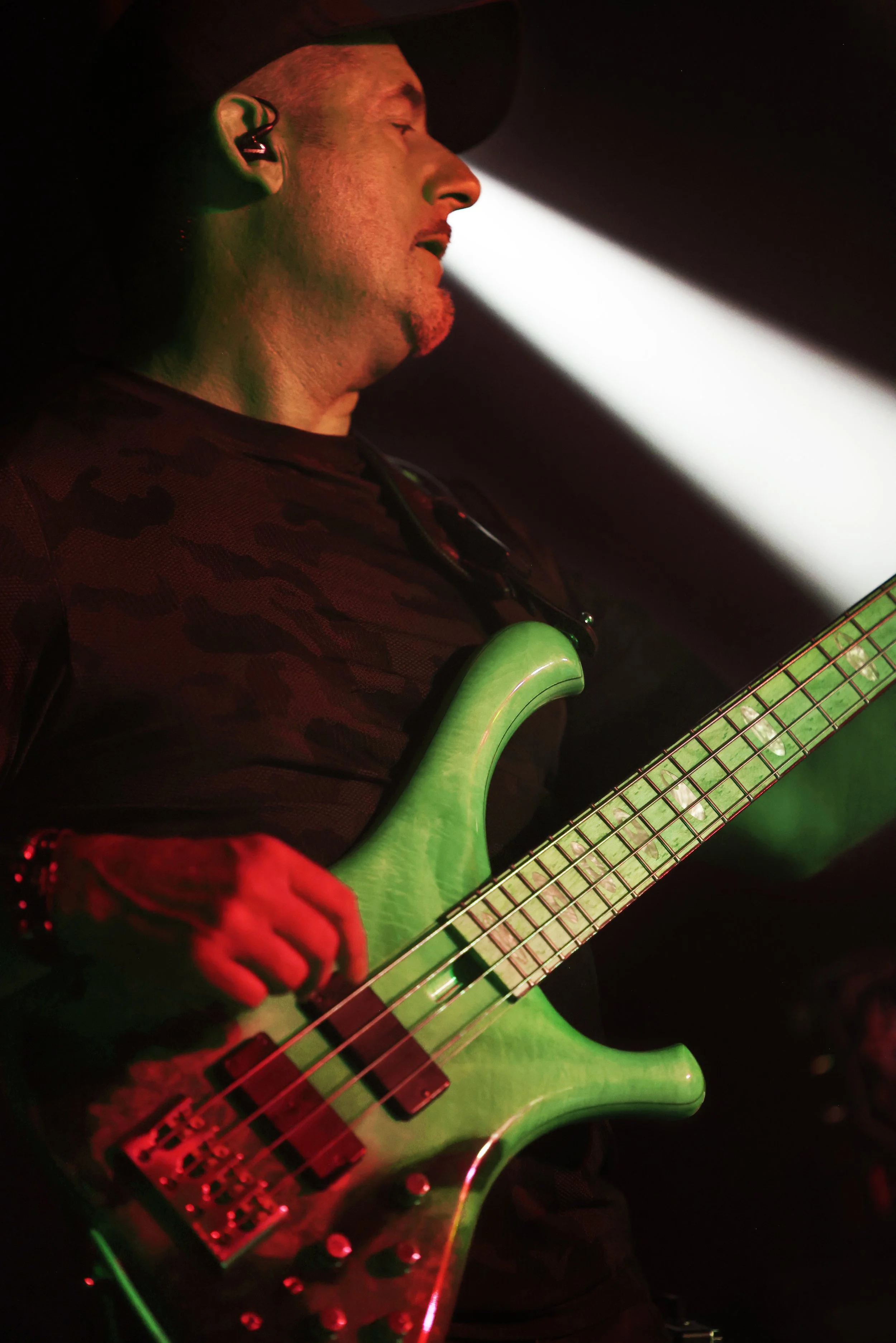 A musician playing a bass guitar on stage, illuminated by red and green lighting, with a focused expression.