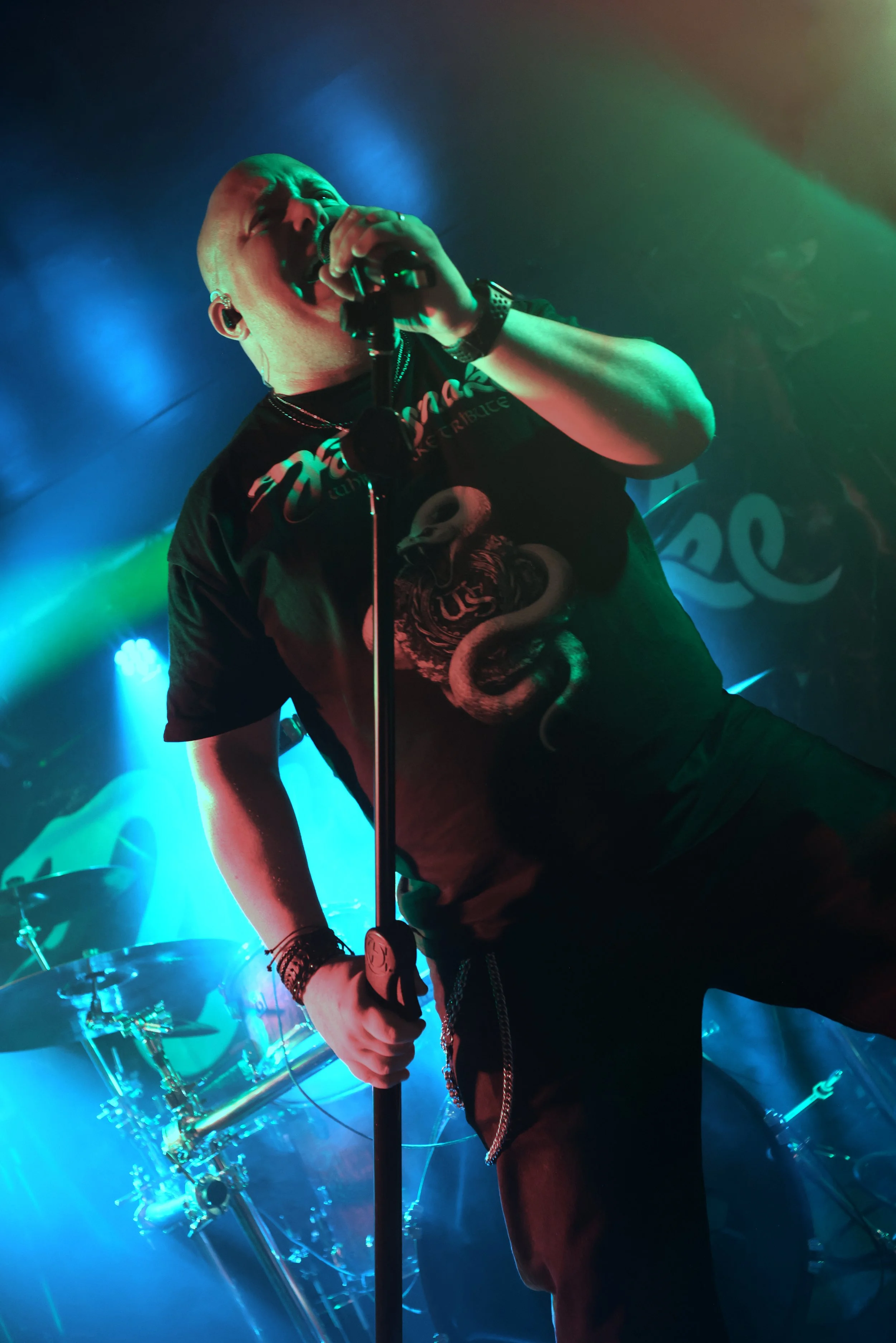 A man singing into a microphone on stage, illuminated by colourful lights, with a drum set visible in the background.