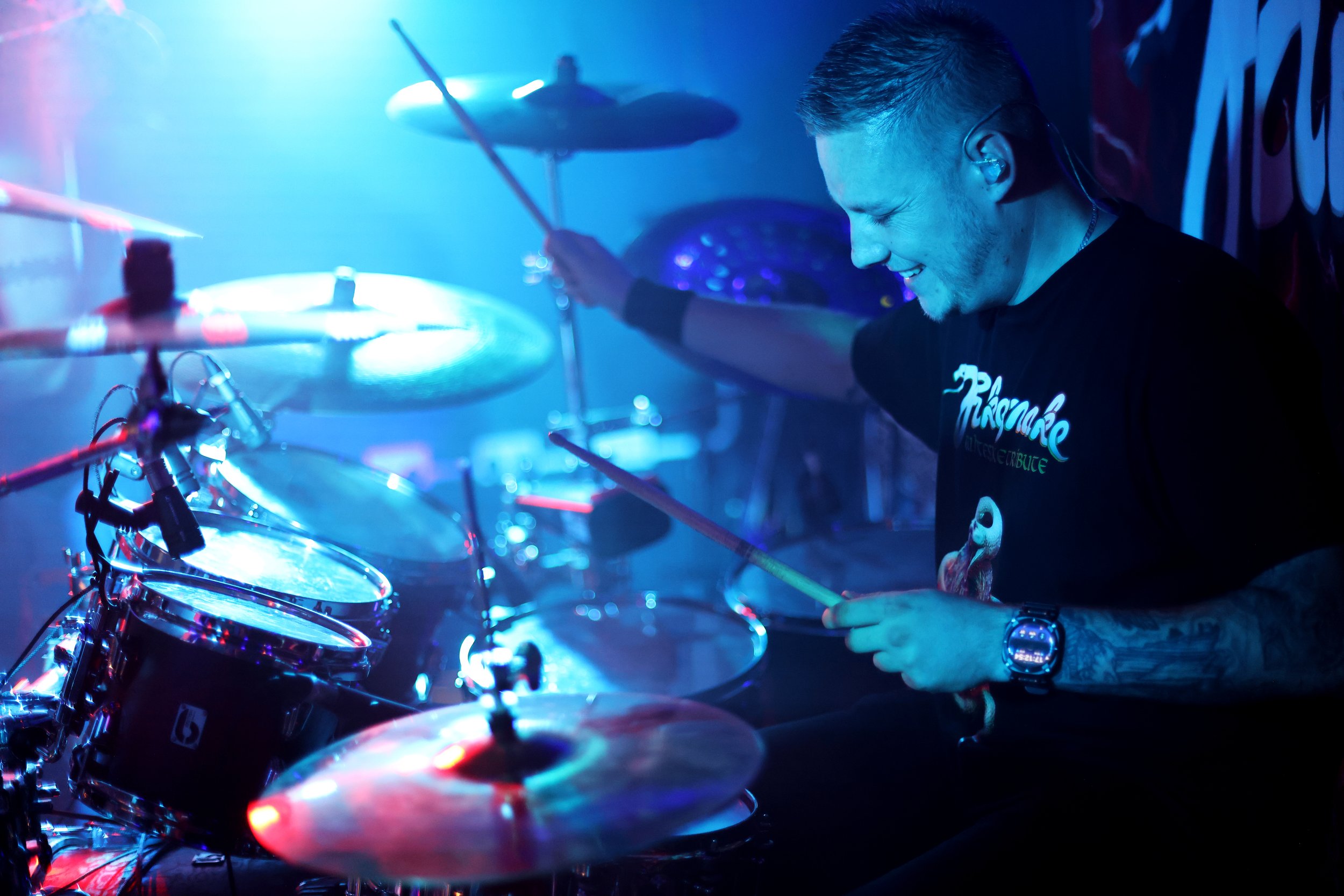 A person with short hair playing a drum set on stage with blue lighting and a smile.
