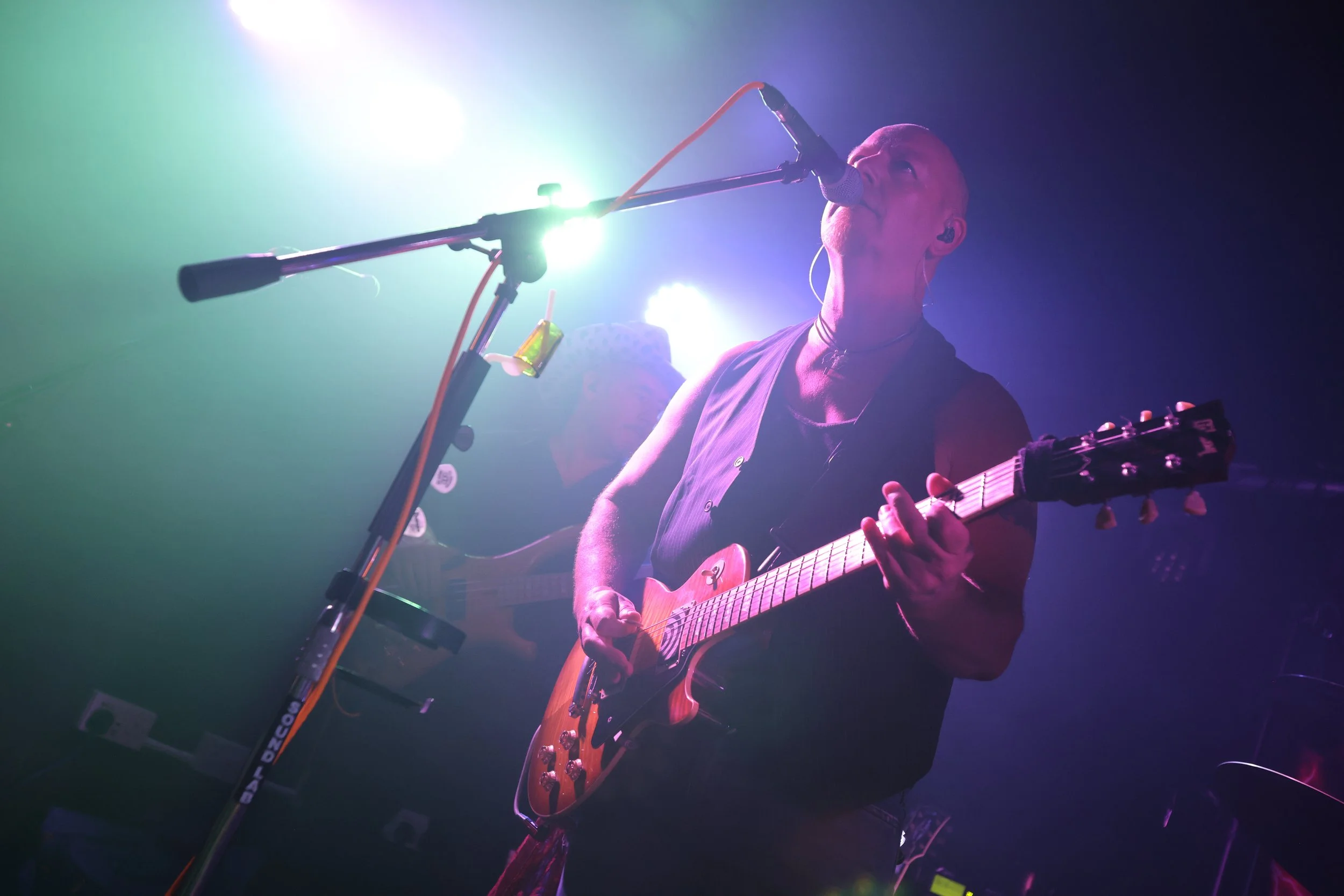 A person playing an electric guitar on stage, singing into a microphone, with colourful stage lights in the background.