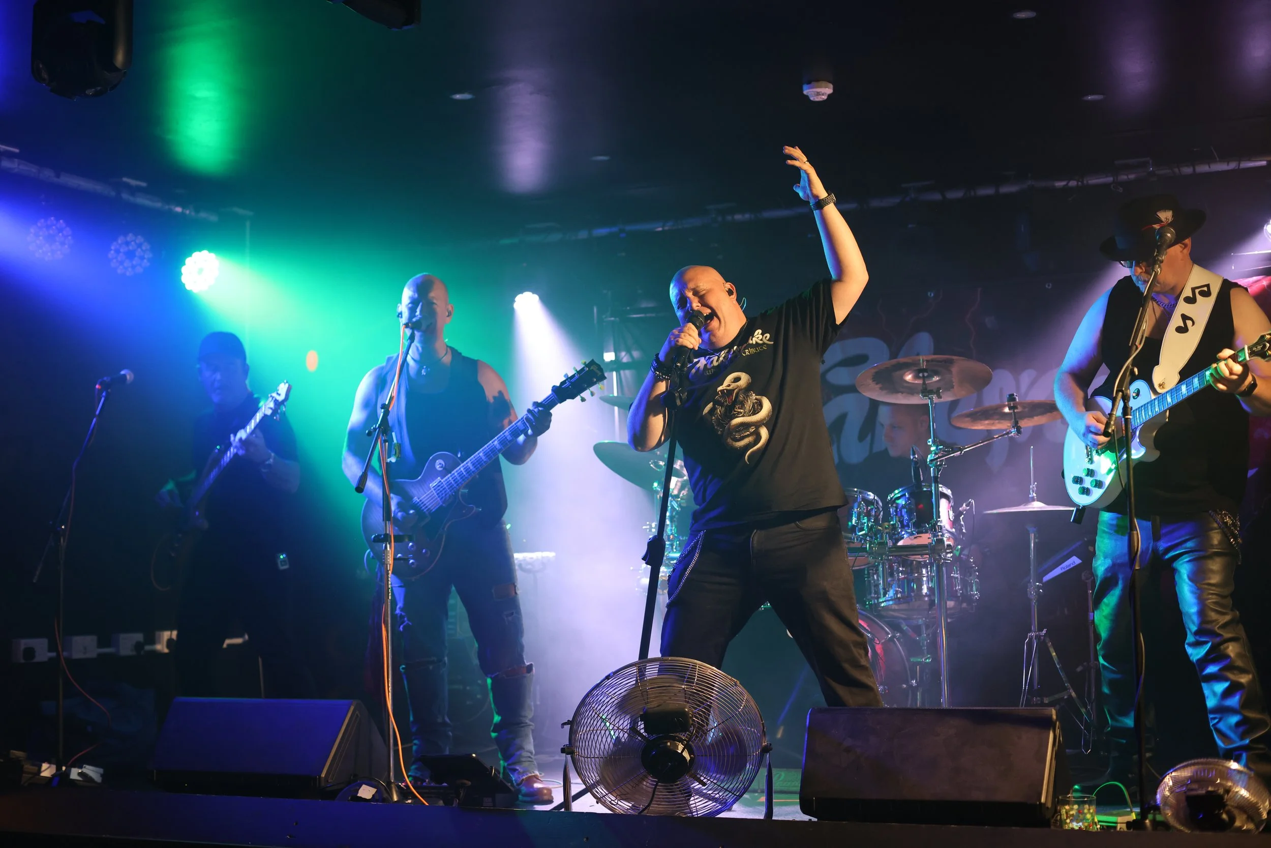 A band performing live on stage with colored stage lights, including a lead singer in the center with a microphone, guitarists on either side, and a drummer in the background.