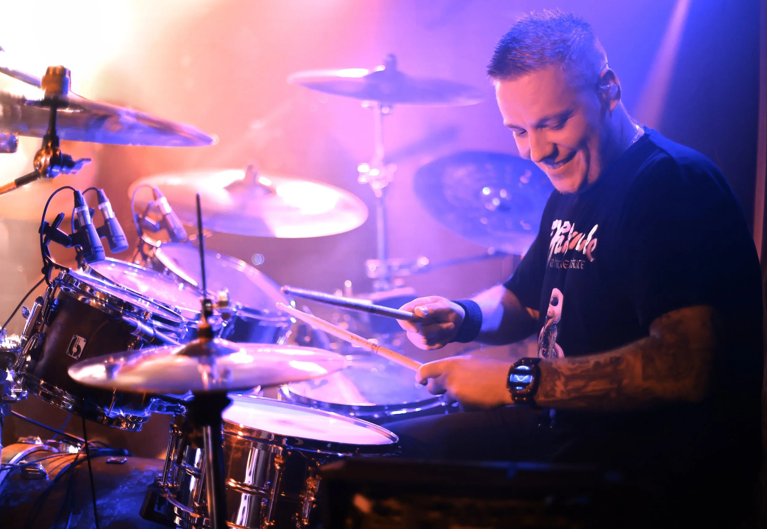 A young man playing the drums on stage, smiling with colourful stage lighting in the background.