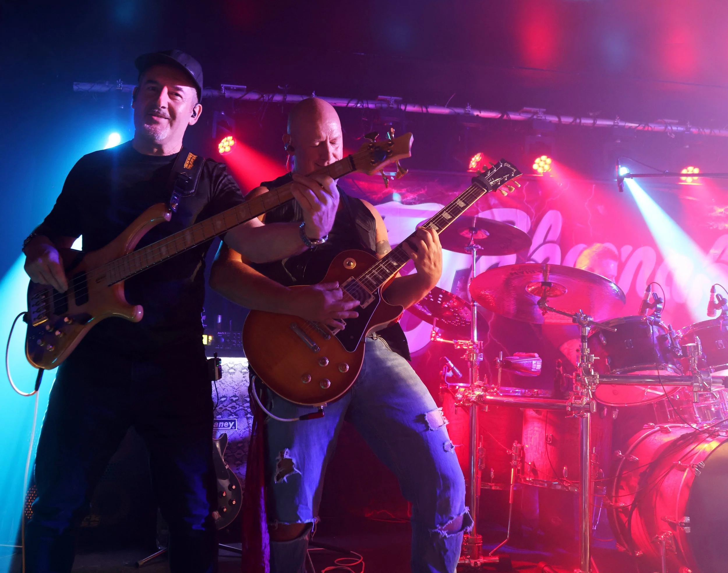 Two musicians playing guitars on stage with colourful lighting and drums in the background.