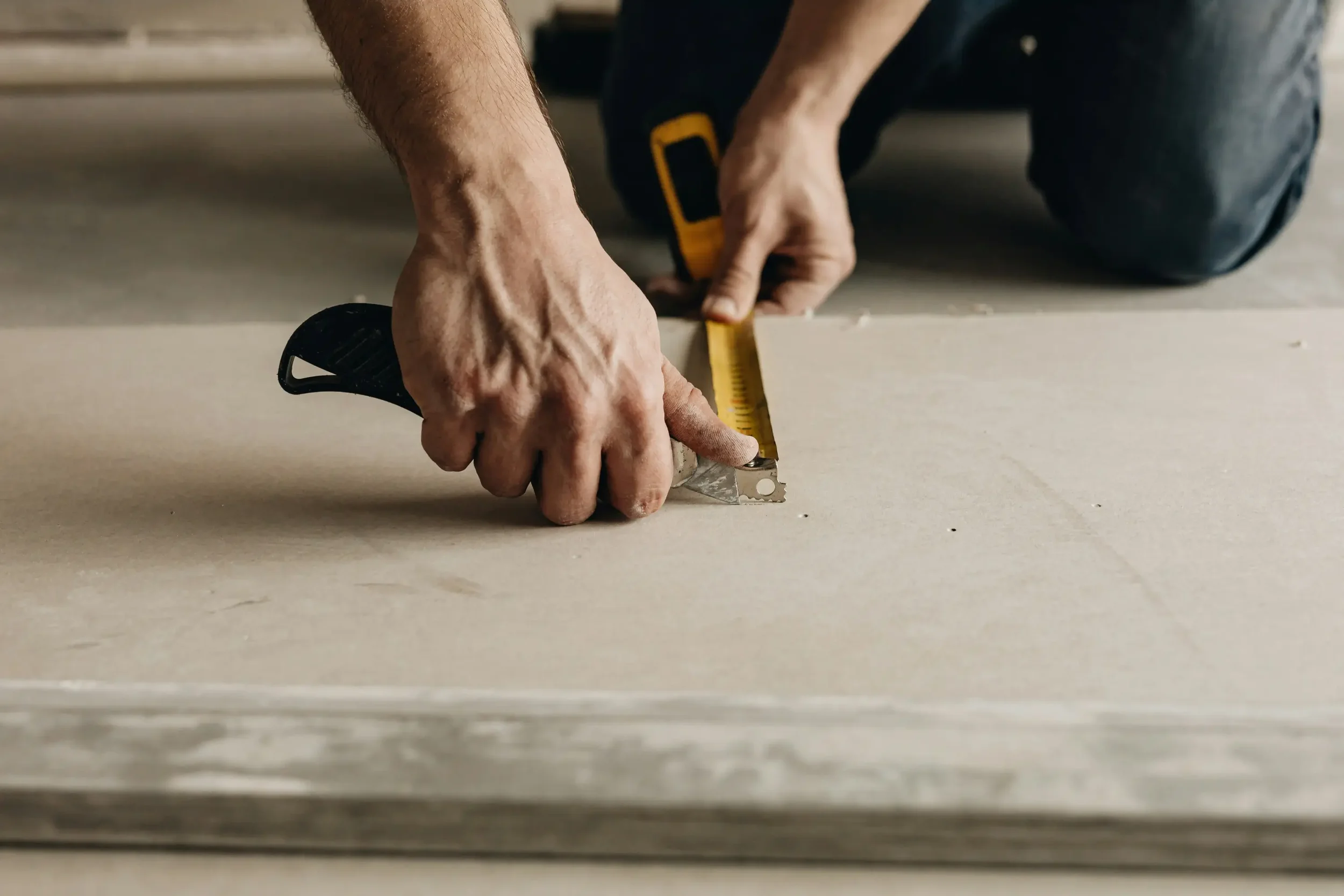 A person is measuring and cutting a piece of wood with a utility knife and a tape measure on a flat surface.