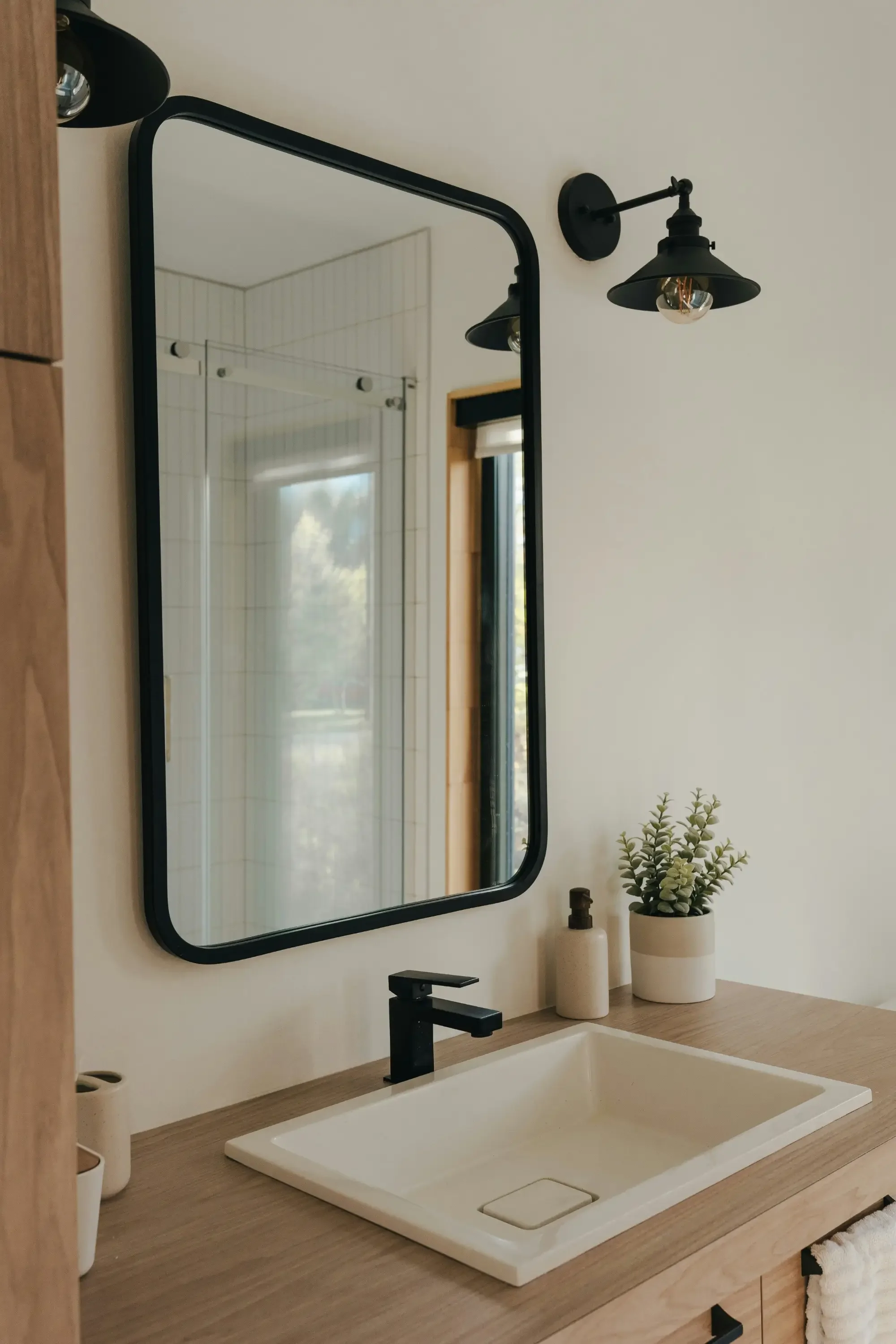Bathroom vanity with a white rectangular sink, black faucet, a round mirror, black wall sconce, potted plant, soap dispenser, and a towel hanging from the cabinet.