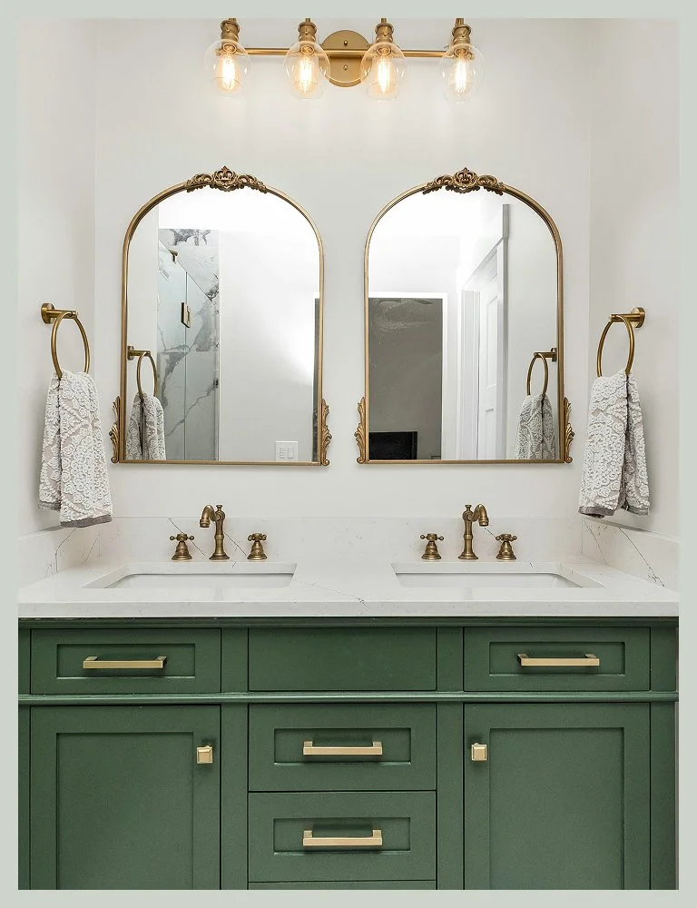 Bathroom with green cabinet vanity, two sinks, and two mirrors with decorative gold frames above each sink. Brass fixtures and towel holders with white textured towels, and a gold light fixture with four exposed bulbs above the mirrors.