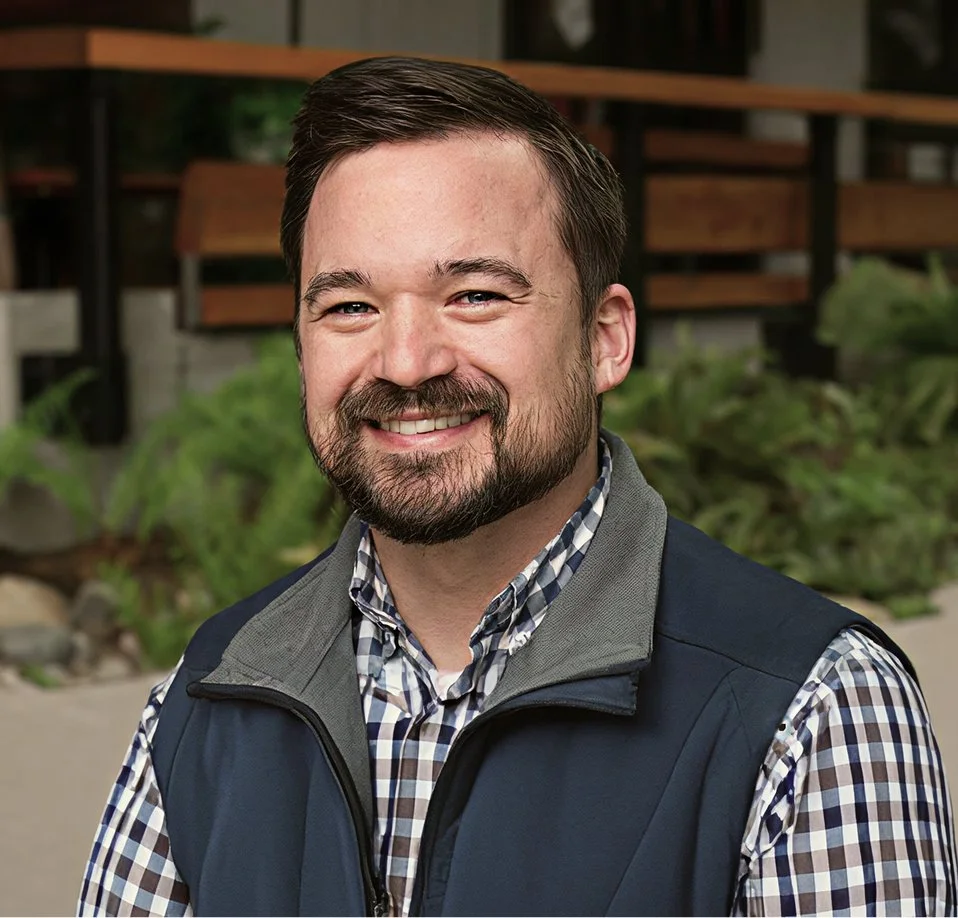 A man with short brown hair and a beard smiling, wearing a checkered shirt and a vest, standing outdoors with greenery and wooden benches in the background.