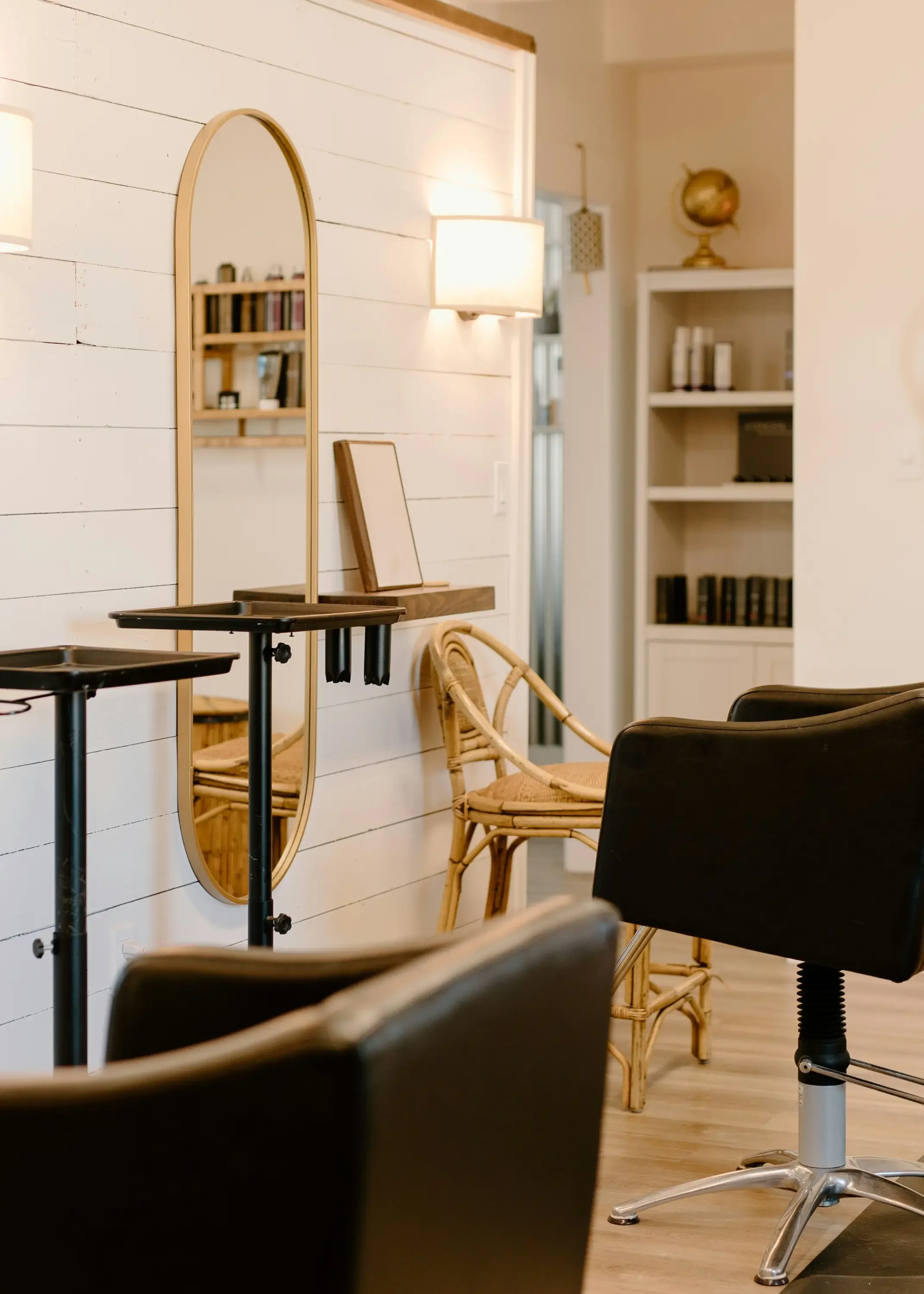Interior view of a room with a wall mirror, a wooden shelf with books, and some chairs including a black office chair and rattan chairs, in a cozy and well-lit space.