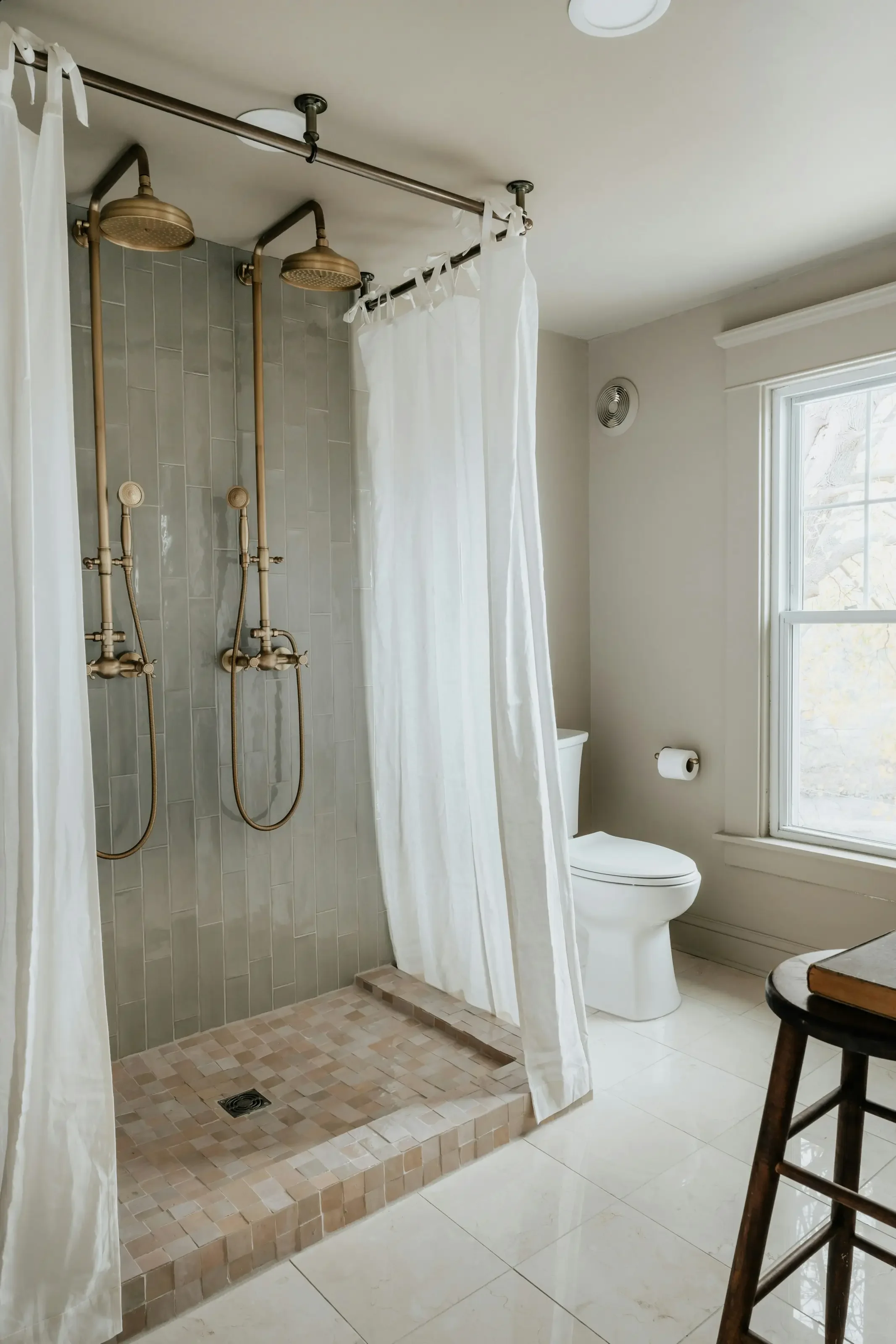 Bathroom with a walk-in shower featuring two vintage-style shower heads, white shower curtain, toilet, large window, and a wooden stool.
