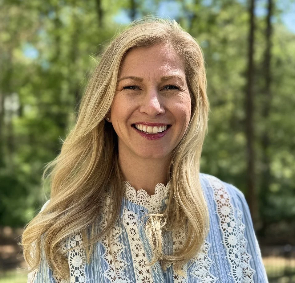A woman with long blonde hair smiling outdoors with trees in the background.