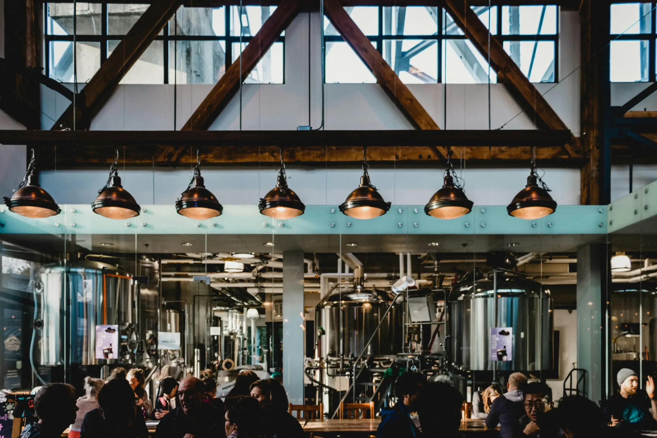 Interior of a brewery with large stainless steel tanks, industrial lighting, and people sitting at tables.