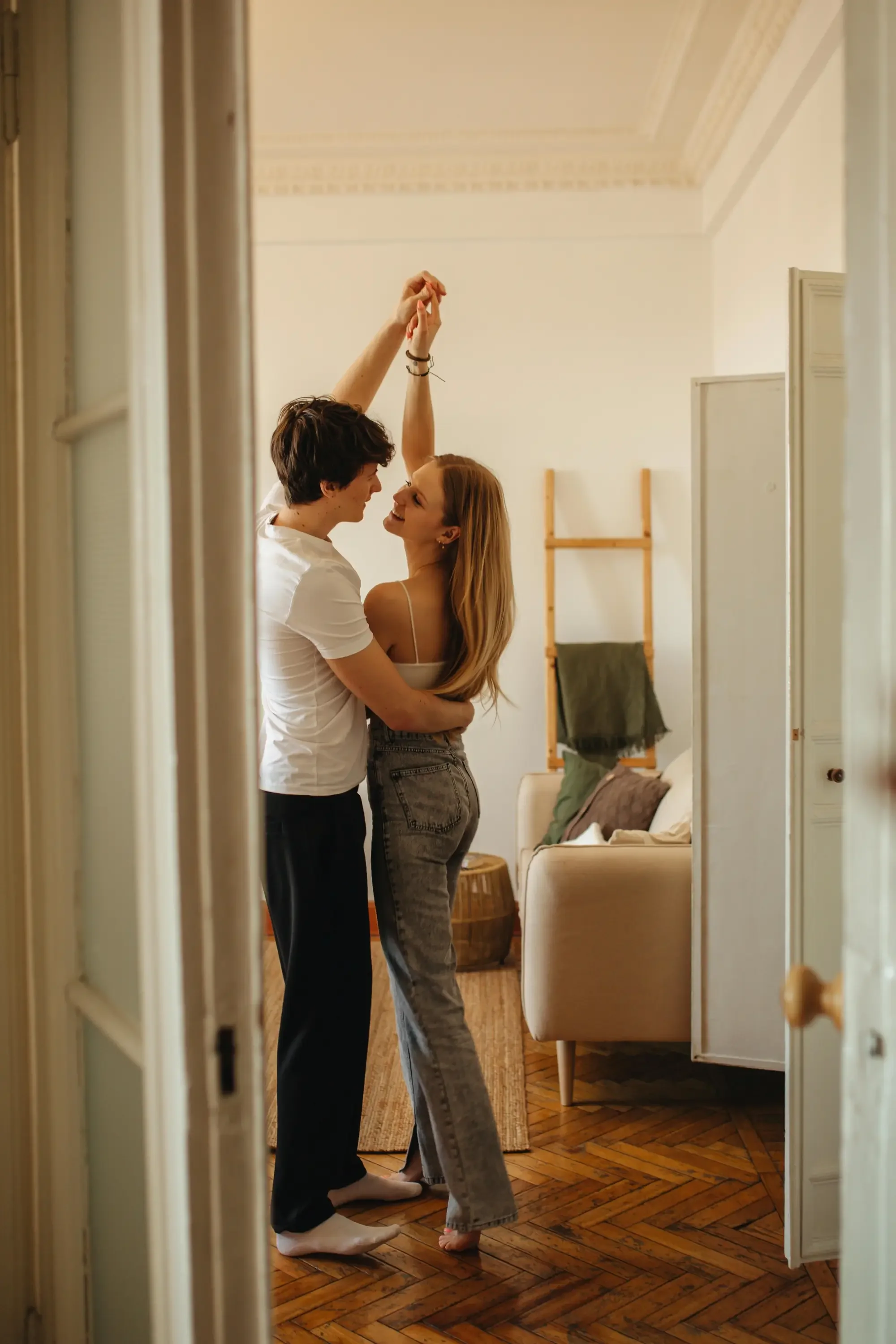 A young couple dancing and smiling in a cozy, well-lit room with hardwood floors and casual decor.