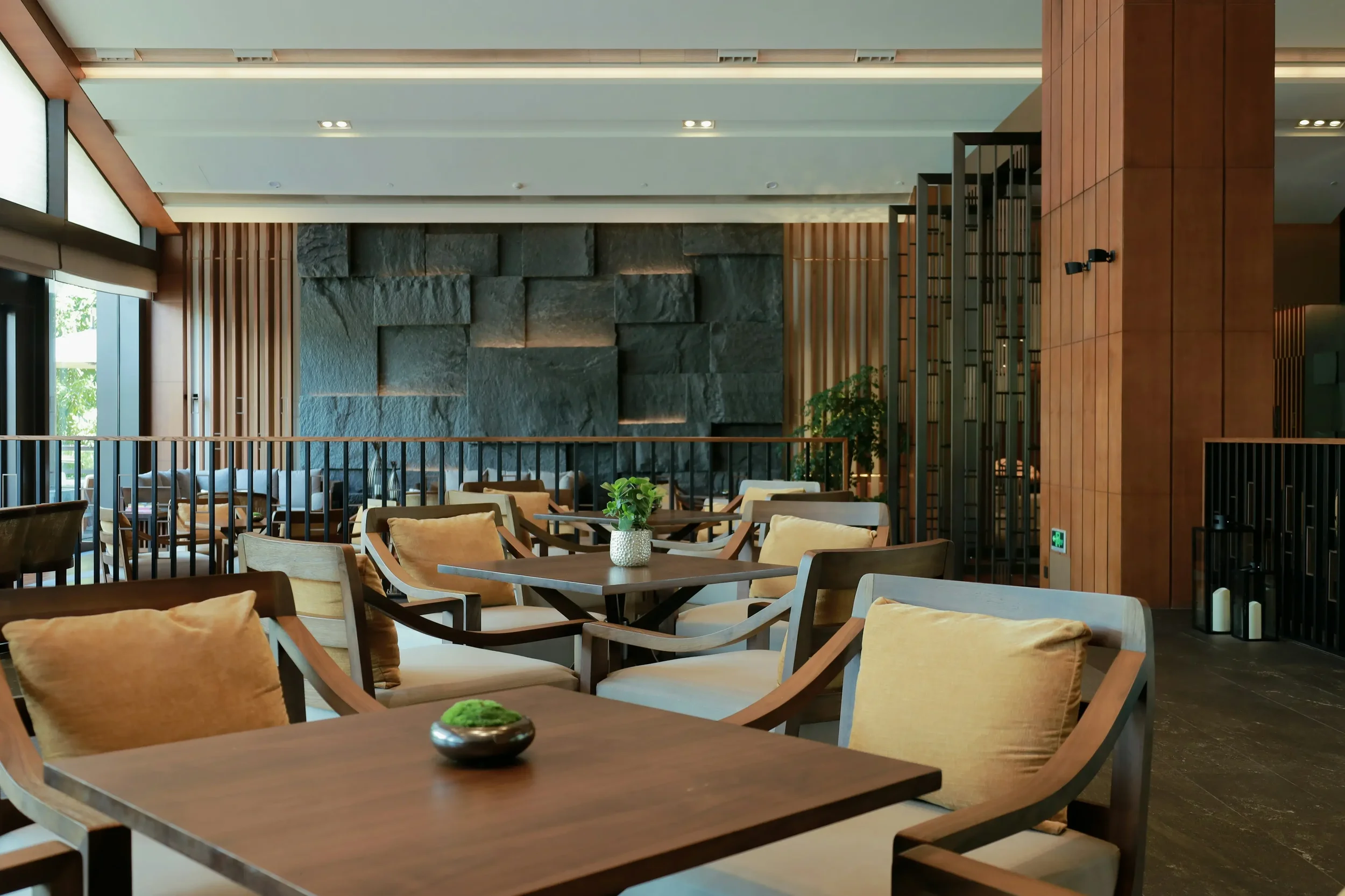 Interior of a modern lounge or restaurant with wooden tables and cushioned chairs, a stone accent wall, and large windows with natural light.