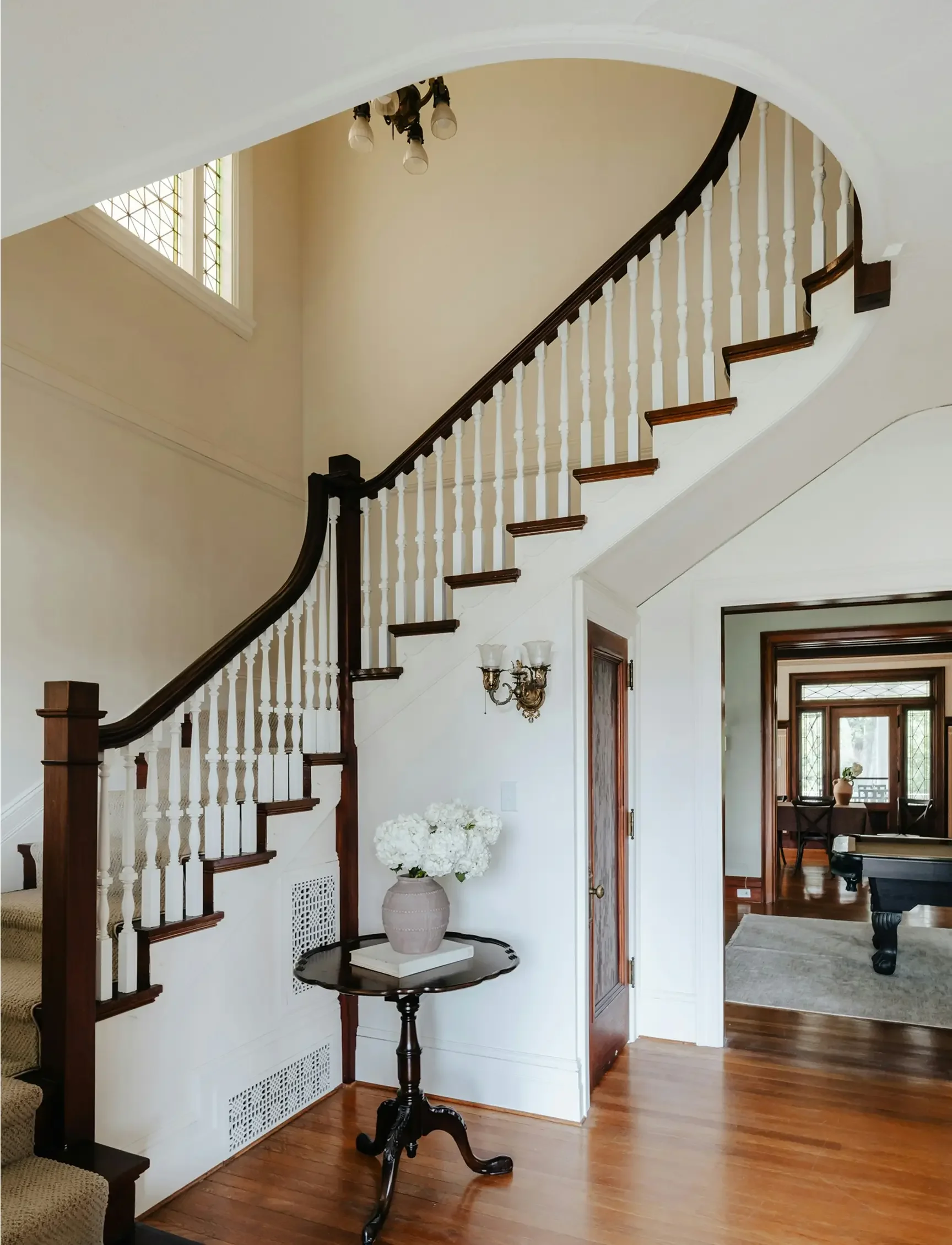 Interior view of a house with a staircase, wooden flooring, a small table with a vase of white flowers, a wall-mounted light fixture, and a hallway leading to a room with large windows.