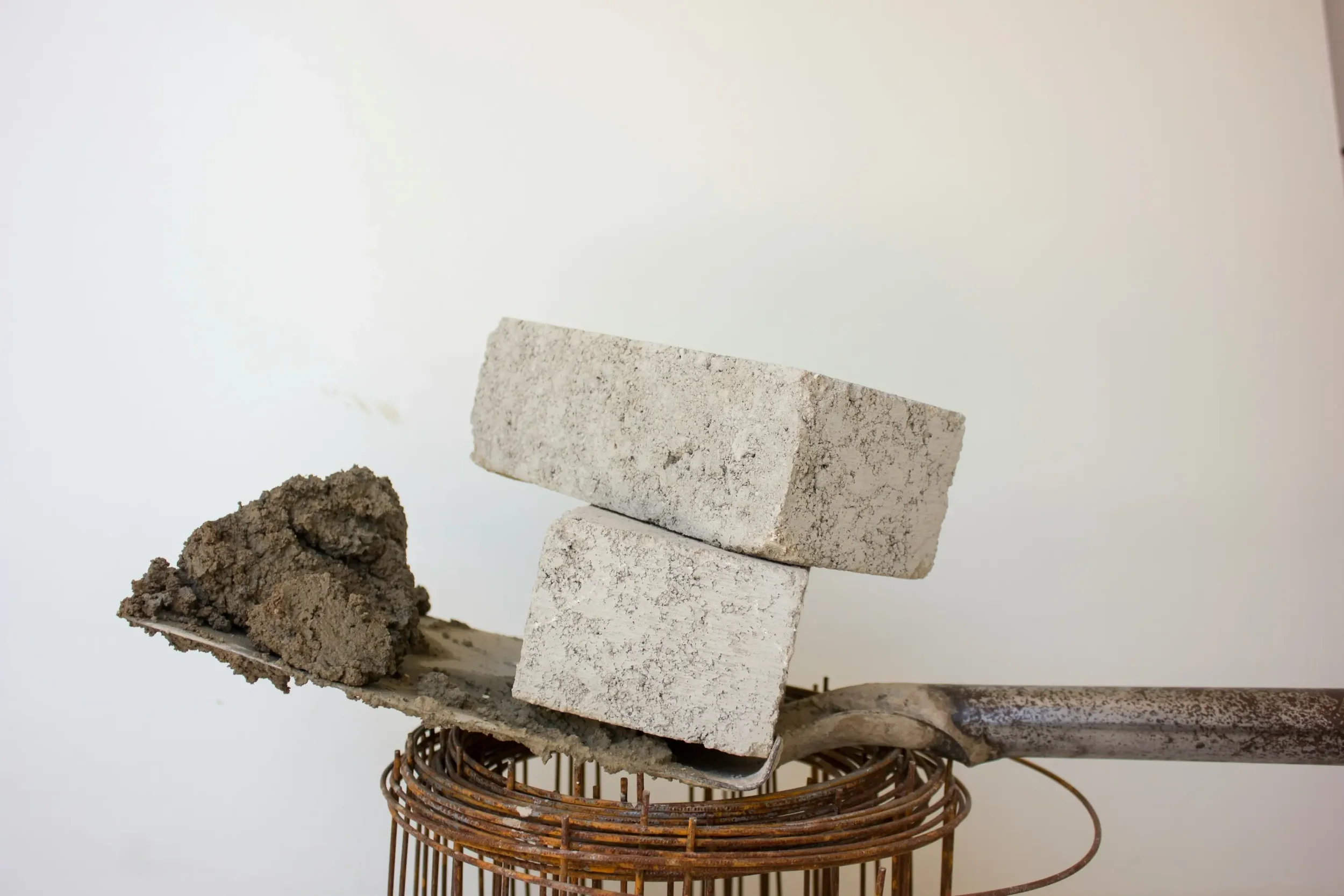 Two white bricks balanced on top of each other, with a weathered metal tool and some dirt or cement on a wire stand against a plain white background.
