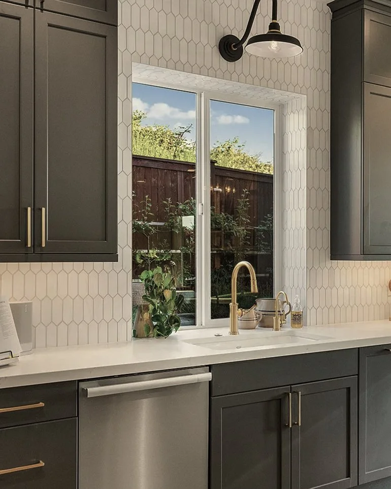 Modern kitchen with gray cabinets, white hexagon tile backsplash, and a white countertop. A window above the sink shows a backyard with a wooden fence and greenery. The sink has a gold faucet and there are small potted plants and soap dispensers on the counter. A black and white wall light fixture is also visible.