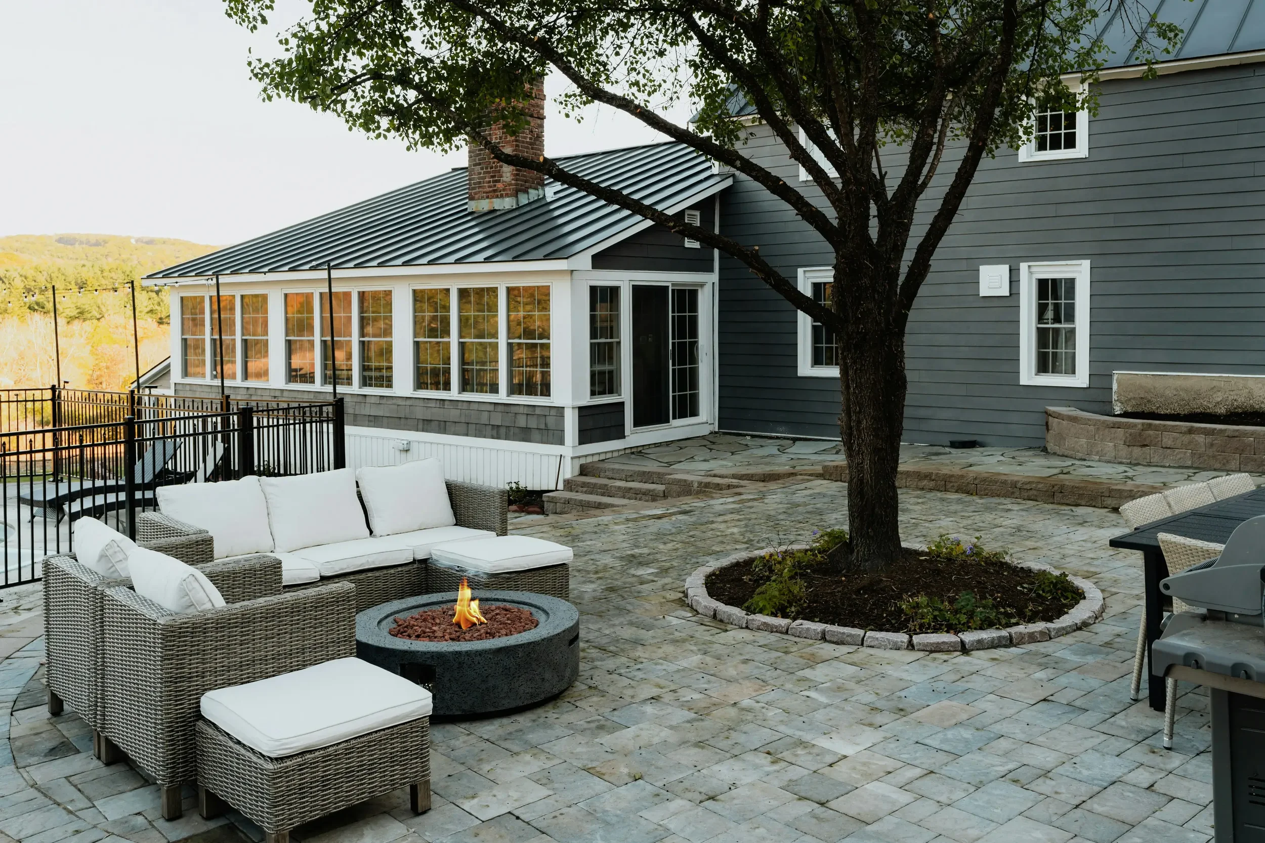 Patio area with outdoor furniture, a fire pit, tree, and a house with a sunroom, in a suburban setting.