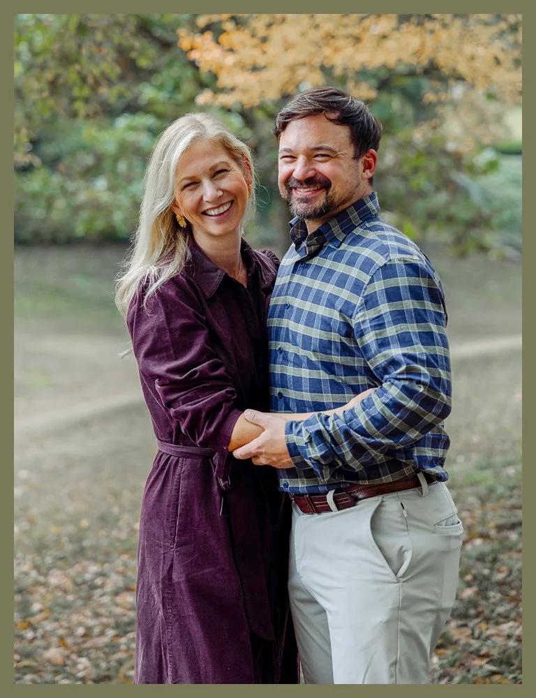 A happy couple standing outdoors in a park during autumn, embracing and smiling at the camera.