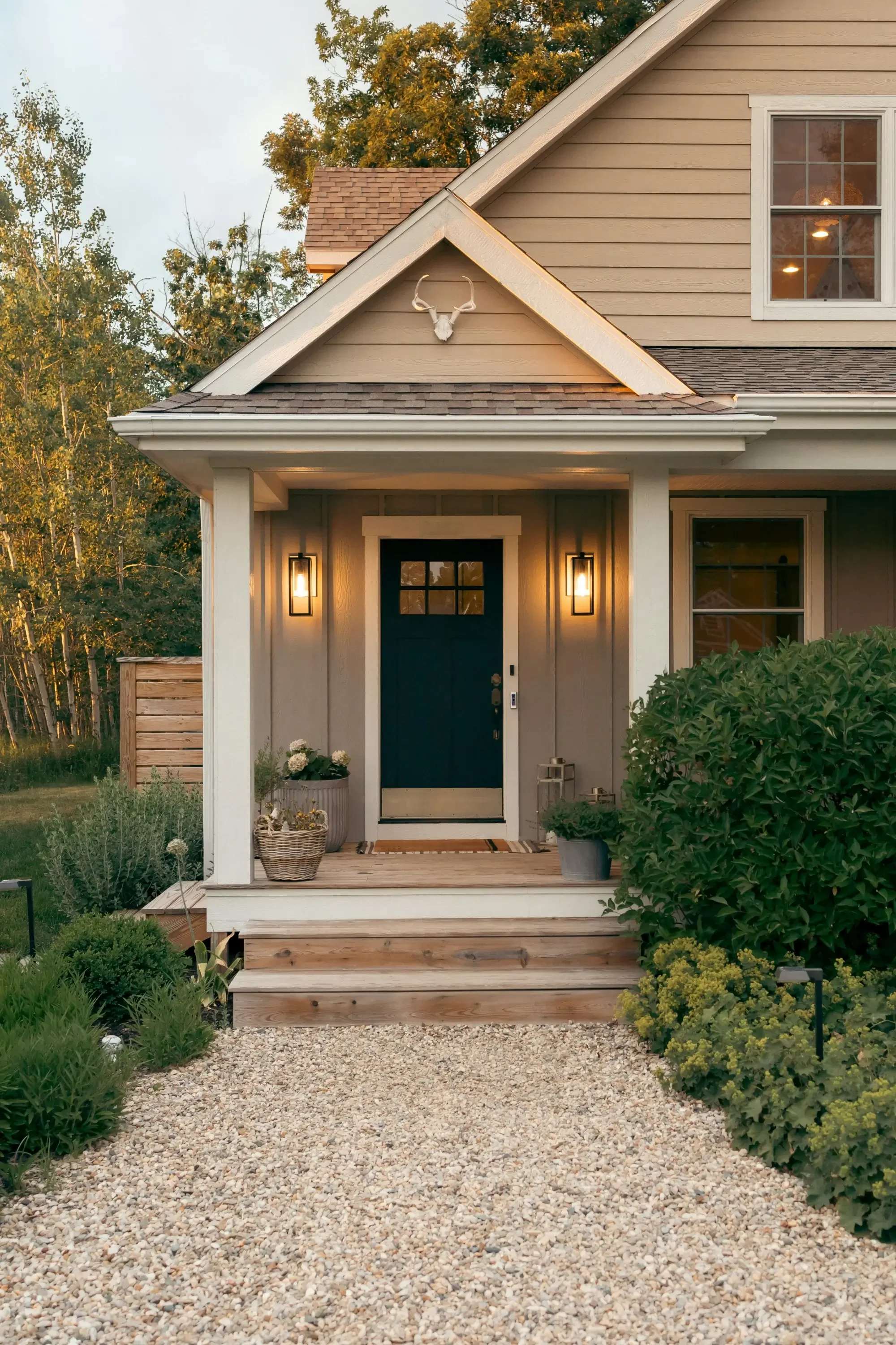 Front porch of a modern house with a dark door, two outdoor wall lights, potted plants, and a gravel pathway leading up to the steps.