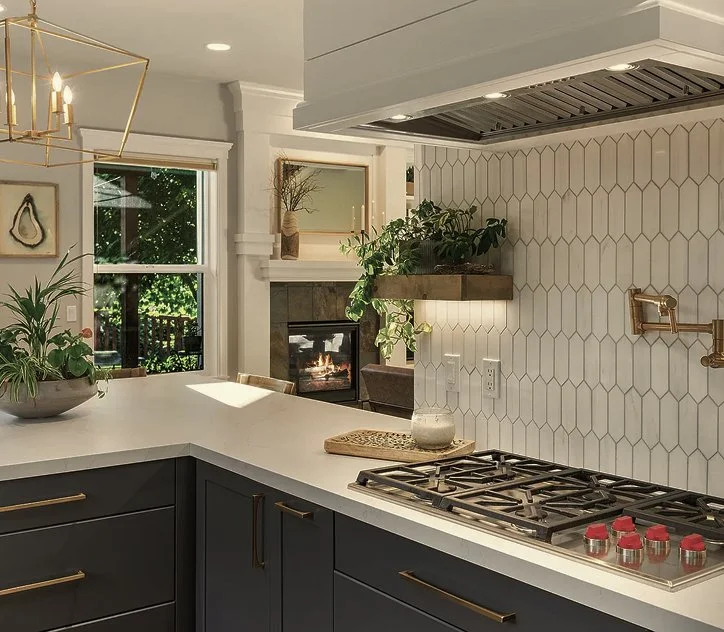 Modern kitchen with dark cabinets, white quartz countertop, and a gas stove. There is a decorative backsplash with a geometric tile pattern, a hanging brass faucet, and green plants. A window and a fireplace are visible in the background.