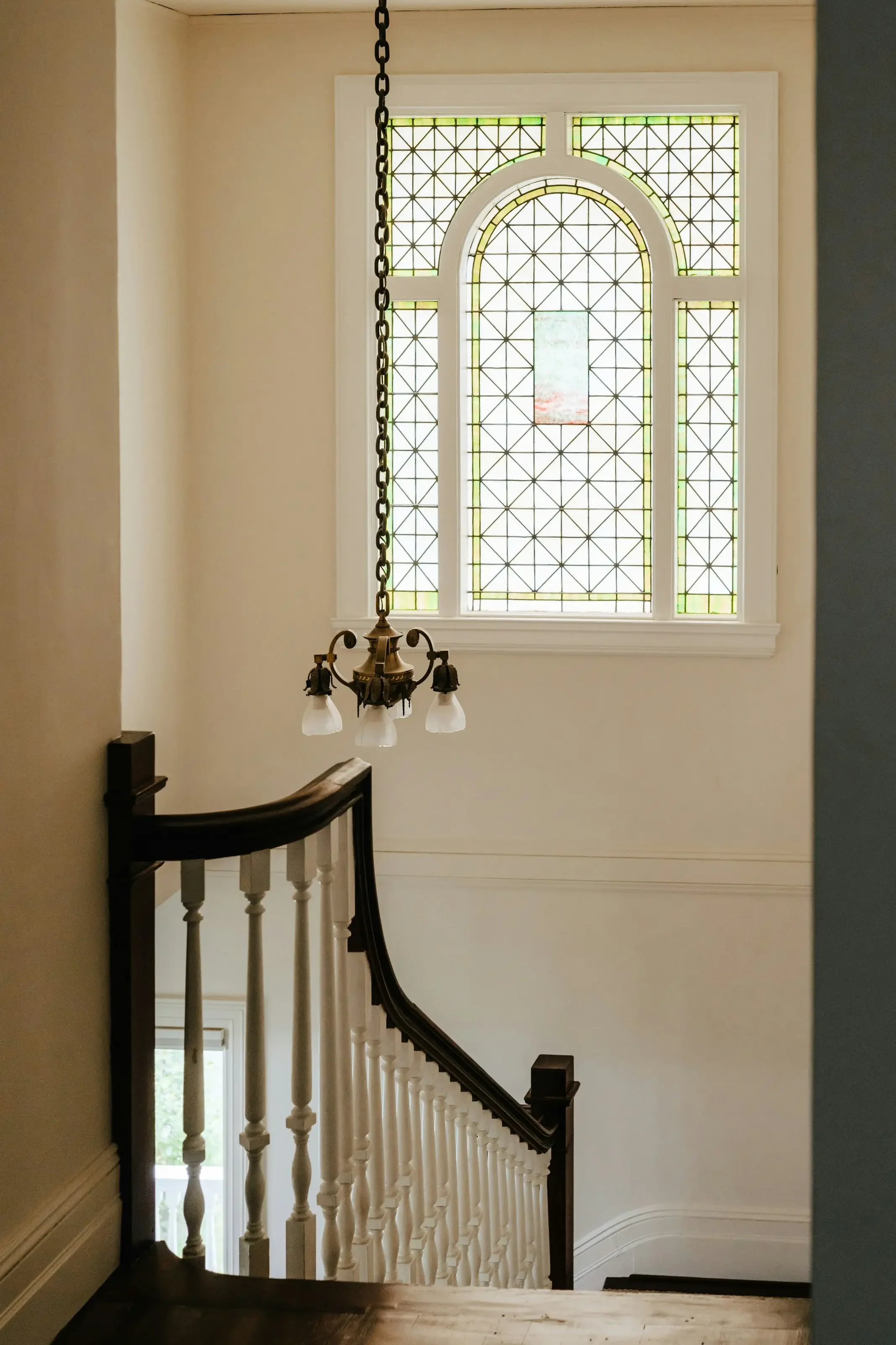 Interior view of a staircase with a window featuring decorative stained glass, a chandelier hanging from the ceiling, and wooden handrails.