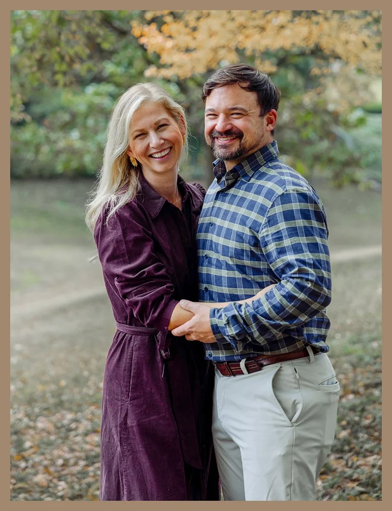 A smiling couple standing outdoors in a park during fall, the woman with long blonde hair wearing a dark purple coat, and the man with dark hair and a beard wearing a blue plaid shirt and khaki pants, holding hands and looking happy.