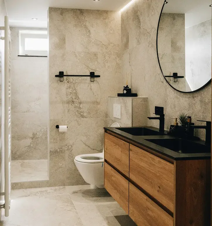 Modern bathroom with a wooden vanity, black sink and faucet, round mirror, toilet, and a small window letting in natural light, with beige tiled walls and floor.