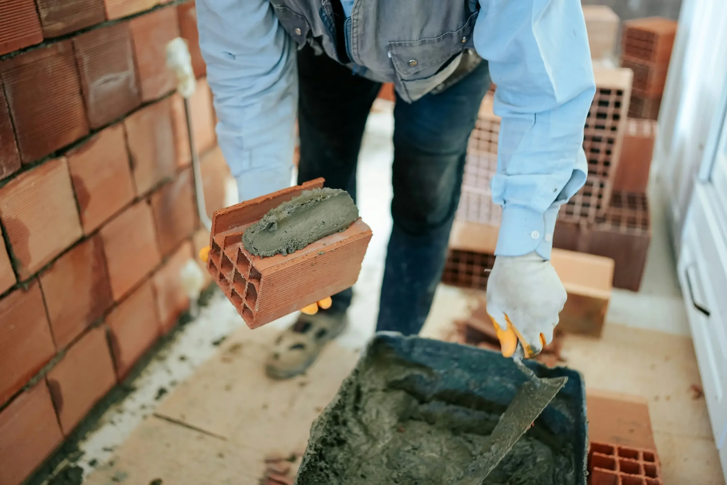 Person laying bricks with mortar in a construction site, with stacked bricks in the background.
