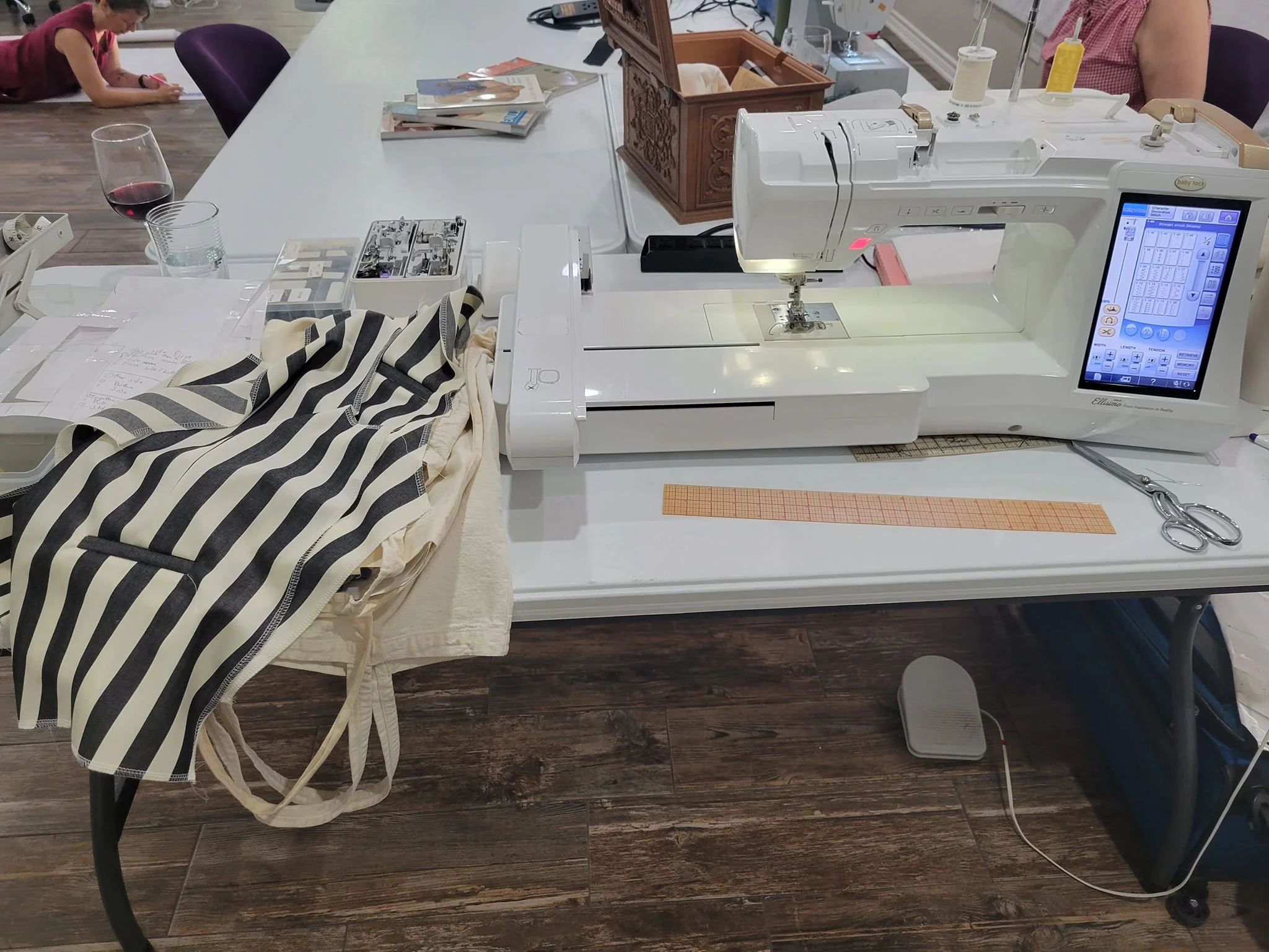 A sewing machine on a table with fabric, scissors, and sewing supplies, with a person sewing and another person working in the background.