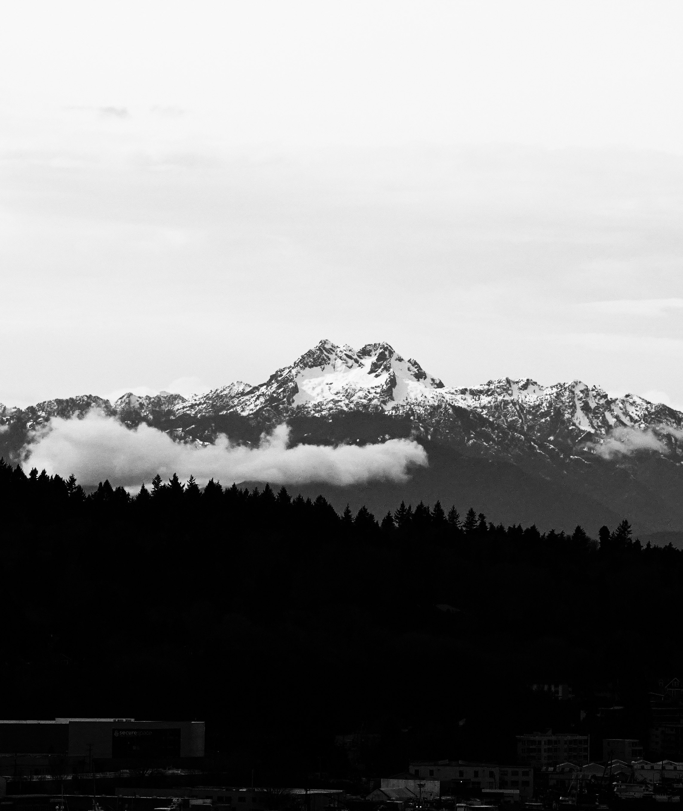 Black and white photograph of a snow-capped mountain range with clouds near the peaks, a forested hillside, and a small town at the bottom.