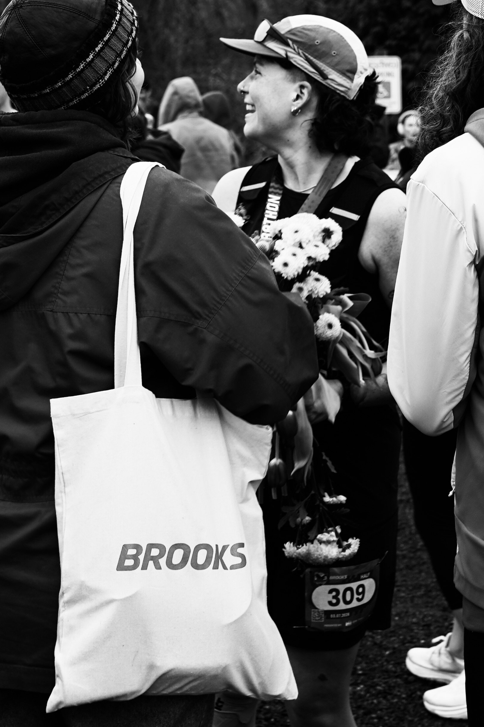 A woman at a marathon event holding flowers and wearing a race bib, talking to another woman with a tote bag that says 'BROOKS' on it.