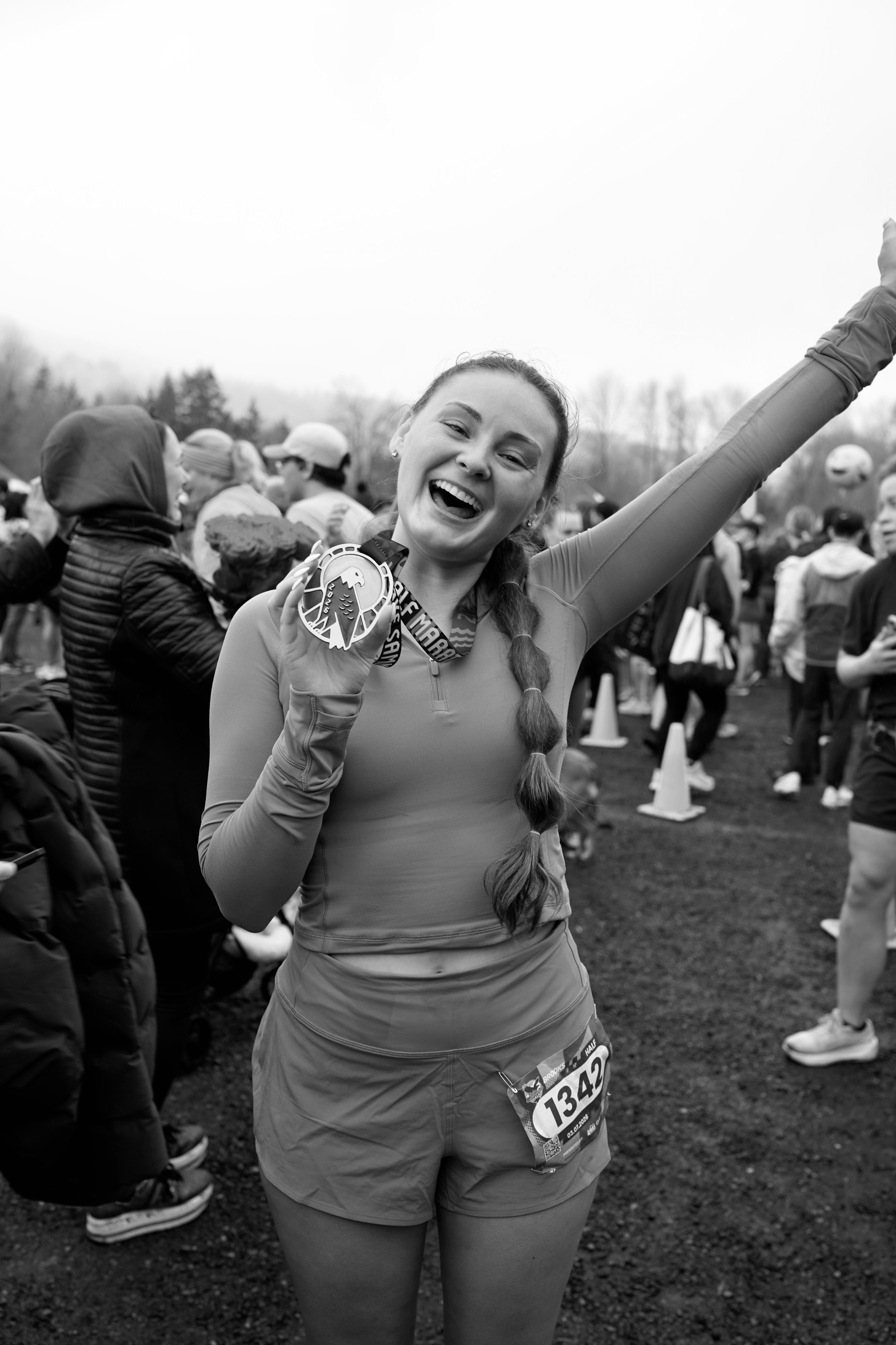 A woman with a long braid, wearing athletic clothing and a race bib, is smiling and holding a medal at a race event with other participants and spectators in the background.